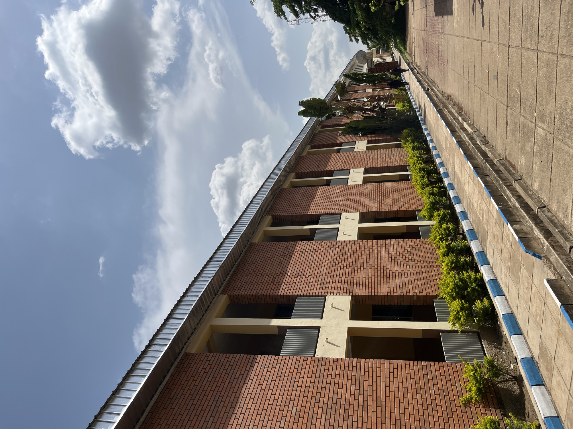 The image shows a brick building with a corrugated metal roof, typical of modern construction in Jos, Nigeria. The building has several windows and balconies, with yellow accents around the openings. A paved walkway runs alongside the building, bordered by a curb painted with alternating blue and white stripes. Small, green shrubs are planted between the curb and the building. Further down the walkway, two individuals, dressed in dark clothing, are walking away from the camera. The sky is partly cloudy with fluffy white clouds against a bright blue background. To the right of the building, there are tall trees, including palm trees, and in the distance, a radio tower is visible. The overall scene suggests an institutional or educational setting within an urban environment.