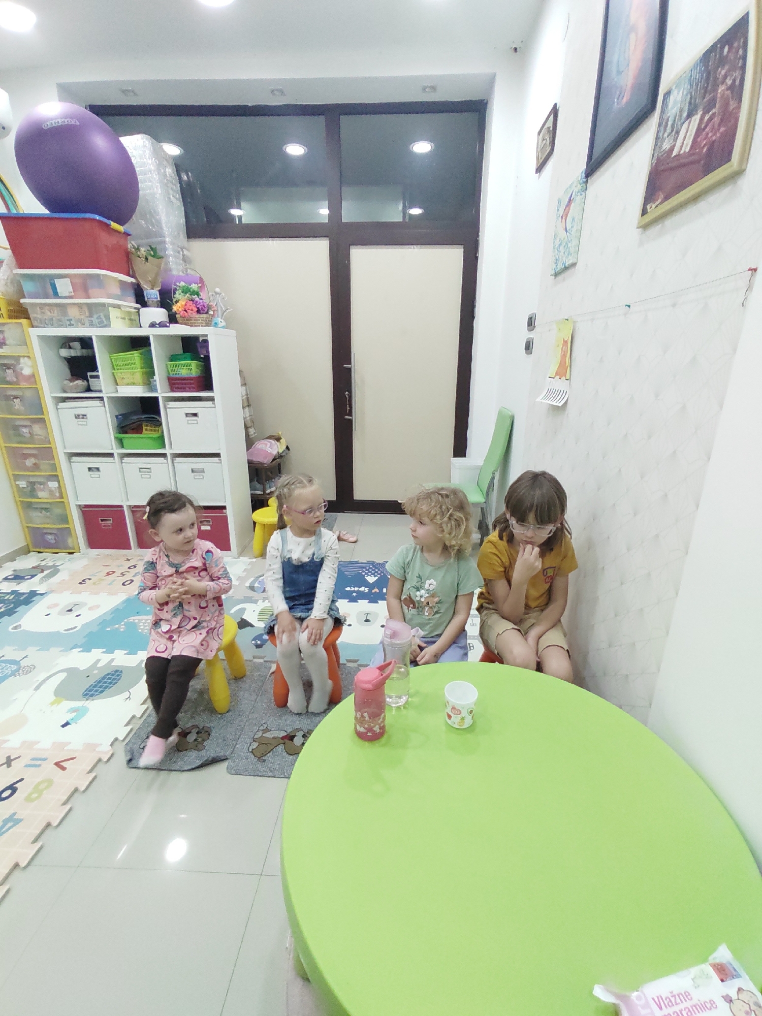 A bright, child-friendly room in what appears to be a preschool or daycare setting. Four young children are seated around a vibrant green, oval-shaped table. A girl wearing a pink patterned dress and brown leggings sits on a yellow stool, another girl wearing a denim dress over a white shirt sits on an orange stool. Two additional children are seated nearby: a girl in a green shirt and a girl wearing an orange top and brown shorts. Several drinking containers are visible on the table. Behind the children, there is a play area with a colorful alphabet-themed floor mat, shelves filled with toys and storage containers. A large purple exercise ball is stacked on top of shelves. Artwork and posters adorn the walls. The interior is in Podgorica, Montenegro.