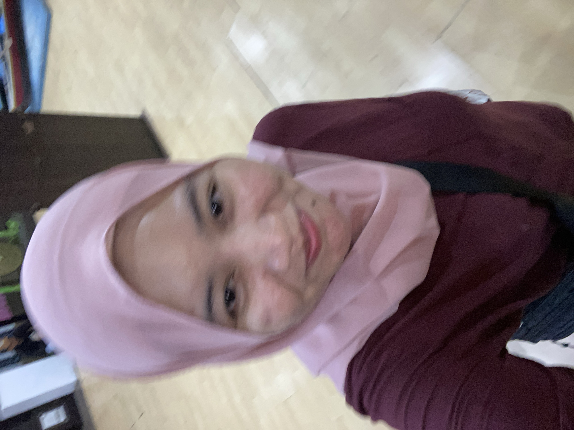 A selfie is taken indoors, focusing on the upper body of a woman wearing a pink hijab and a maroon shirt. She is smiling at the camera. The background is blurred, but appears to be a room with light-colored flooring and some dark vertical structures. A red and white object with a blue base is partially visible in the upper right corner. The scene suggests a casual setting, possibly a public indoor space. The woman is likely in Banjarmasin, Indonesia, based on the provided context.