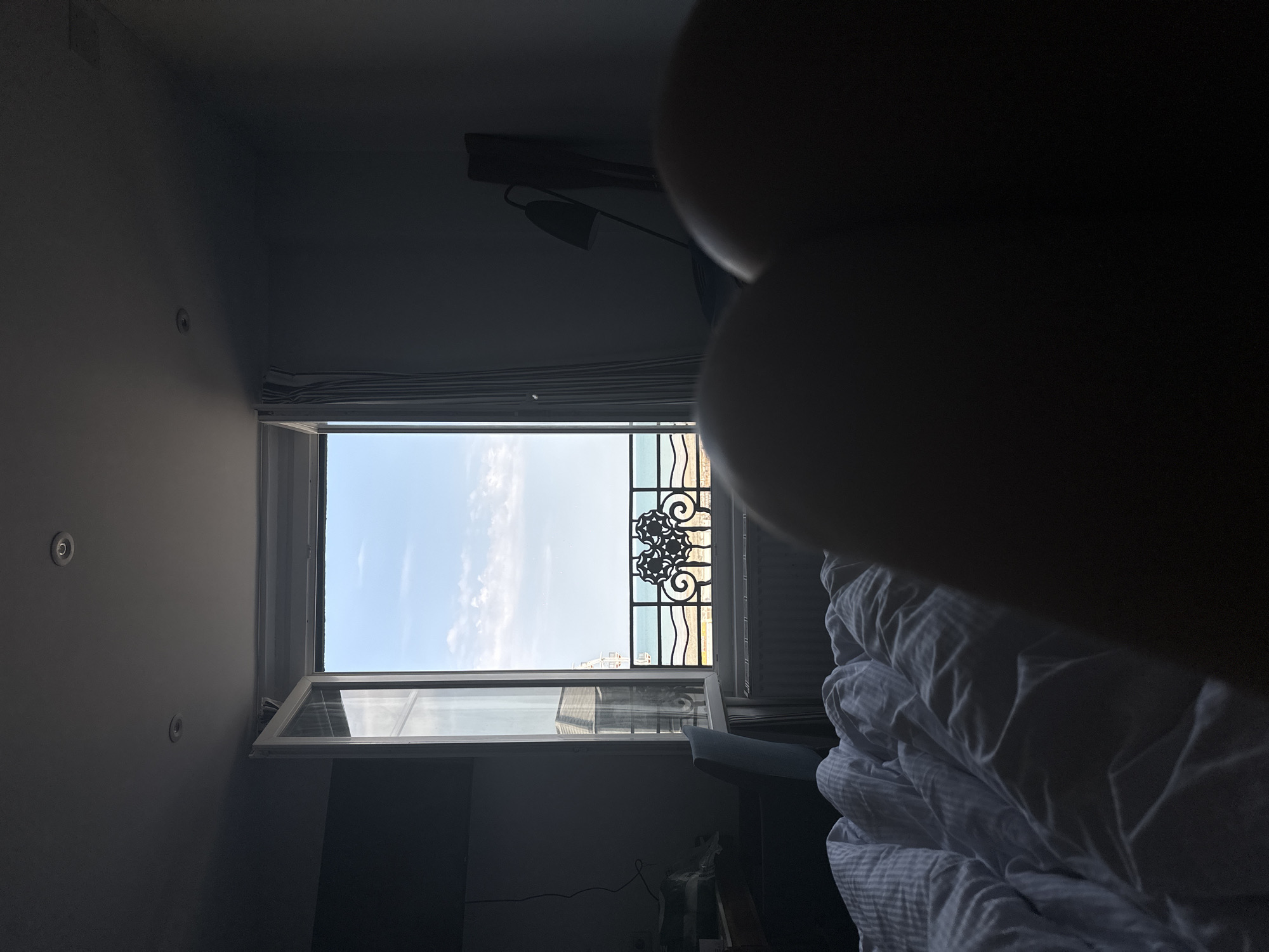 The image shows a bedroom with a window open to a view of the sea and sky in Le Tréport, France. In the foreground, a rumpled white duvet covers a bed. Two dark, out-of-focus shapes, likely parts of a person's body, are positioned in the lower right corner. The window, partially open, reveals a wrought-iron balcony railing with decorative scrollwork. Beyond the railing, a strip of yellow sand is visible, followed by the blue sea and a light blue sky with scattered white clouds. A building with windows and a pale facade is partially visible through the window on the left. To the left of the window, a dark desk with some items on it and a light blue chair are visible. To the right of the window, a dark curtain hangs, and a dark, silhouetted floor lamp with an angled shade stands near a wall. The room appears to be dimly lit, with light streaming in from the window.