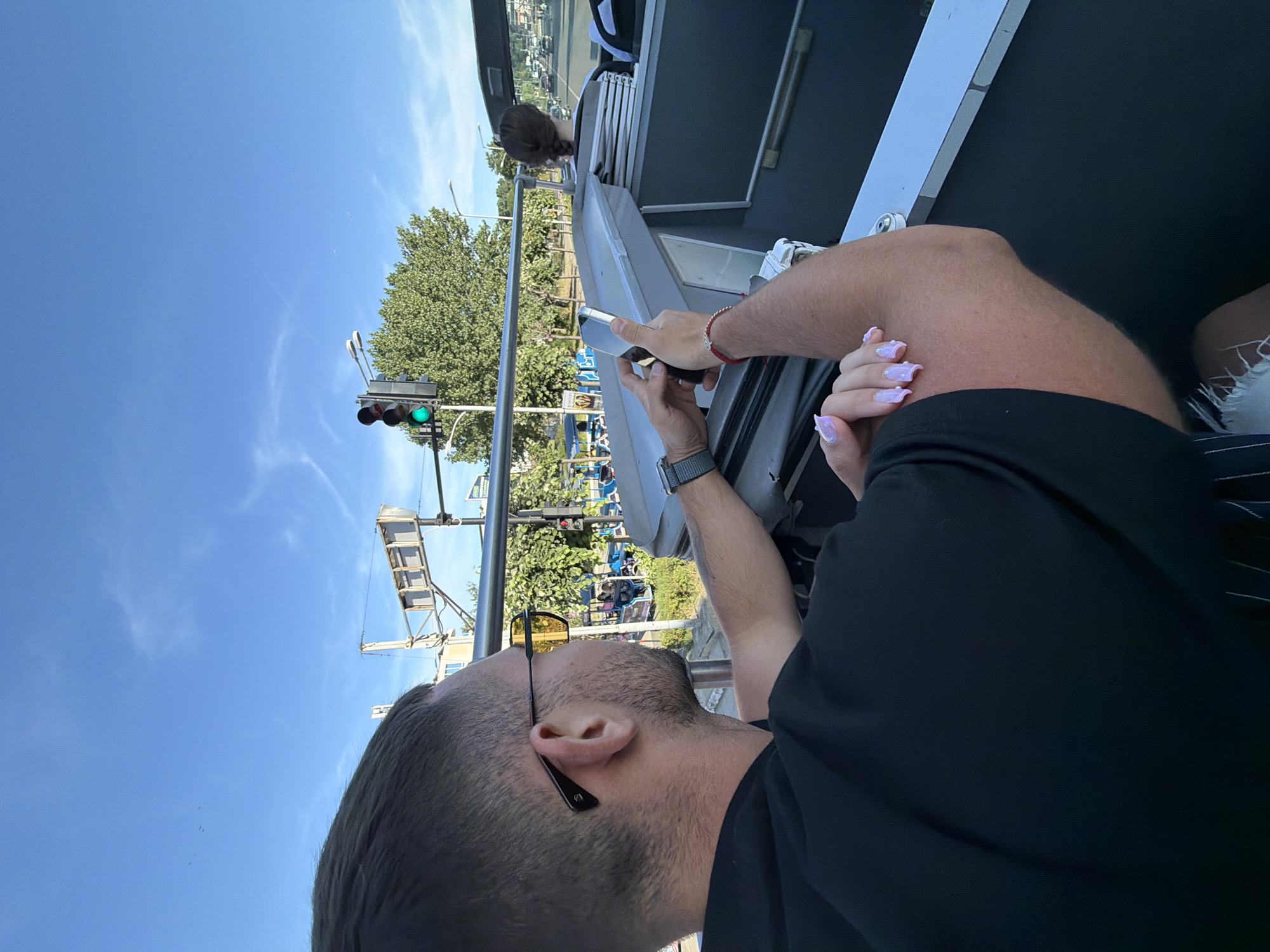 The image captures a bright, sunny day in Nesebar, Bulgaria, likely from the upper deck of a tour bus. In the foreground, a man with short dark hair and sunglasses with yellow lenses is seen from the back, holding a smartphone. His left arm is resting on the bus railing, and his right hand is holding the phone. Next to him, a woman's hand with light purple, pointed nails is visible, suggesting she is also on the bus. In the background, a traffic light with a green signal is prominent, alongside lush green trees. Further back, other people can be seen in what appears to be a public transportation vehicle, and cars are visible on a street, indicating an urban setting. The overall scene depicts a moment of leisure and sightseeing in a Bulgarian city.
