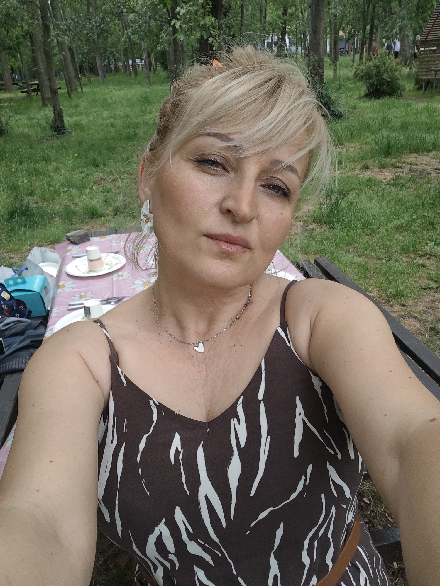 A medium shot captures a mature, fair-skinned woman with blonde hair pulled back in a bun and bangs. She has fair skin, and is wearing a sleeveless brown and white dress with a V-neck and a brown belt. She is also wearing a silver necklace with a heart pendant, and large white flower-shaped earrings.
She is sitting at a picnic table covered with a daisy-patterned pink tablecloth. Dishes, cups, and a blue box are on the table. The background shows a grassy park with trees.
This scene is likely located in a park within the city of Burgas, Bulgaria, based on the environment depicted.