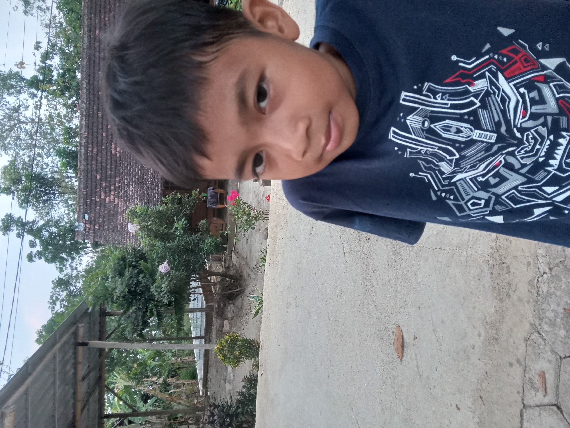 A young boy with dark hair and brown eyes is shown in a close-up shot. He is wearing a navy blue t-shirt with a graphic design on it. The background appears to be an outdoor setting, possibly a rural area in Kedunggudel, Indonesia. There is a building with a tiled roof and some trees visible. The ground is paved with concrete. The lighting suggests it is late afternoon or early evening.