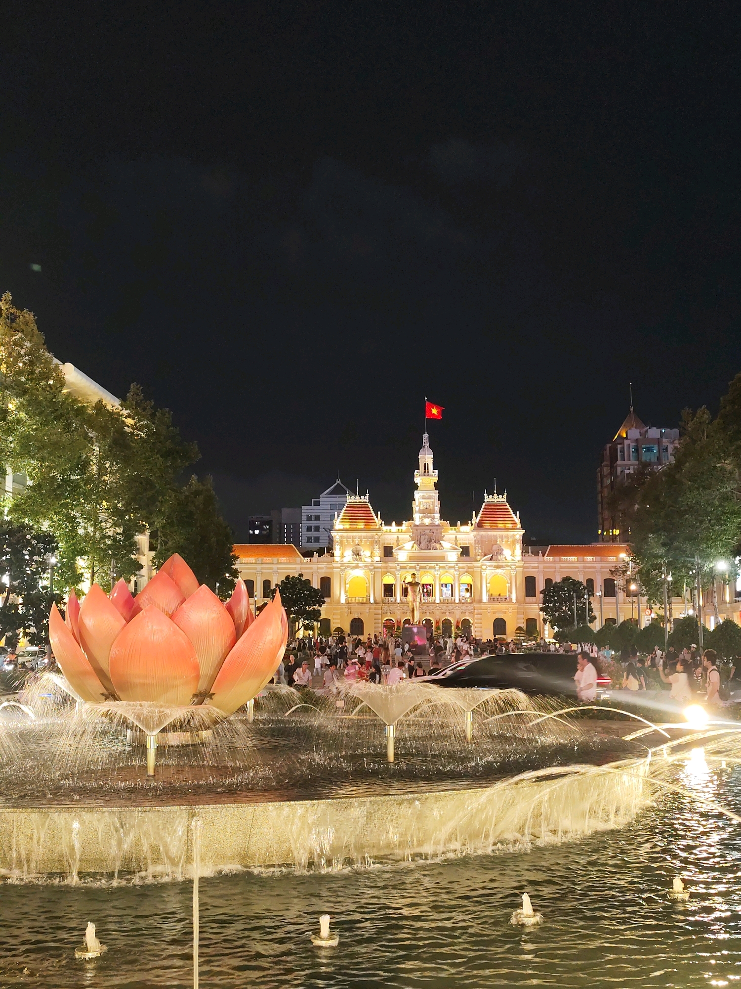 The image shows a night scene of the Saigon City Hall (Hôtel de Ville) in Ho Chi Minh City, Vietnam. In the foreground, a large, illuminated lotus-shaped fountain is active with water spraying. Behind the fountain, a crowd of people is gathered in front of the brightly lit City Hall building. The building features French colonial architecture with a prominent clock tower topped by a Vietnamese flag. Trees and streetlights are visible on either side of the plaza. The sky is dark, indicating nighttime.