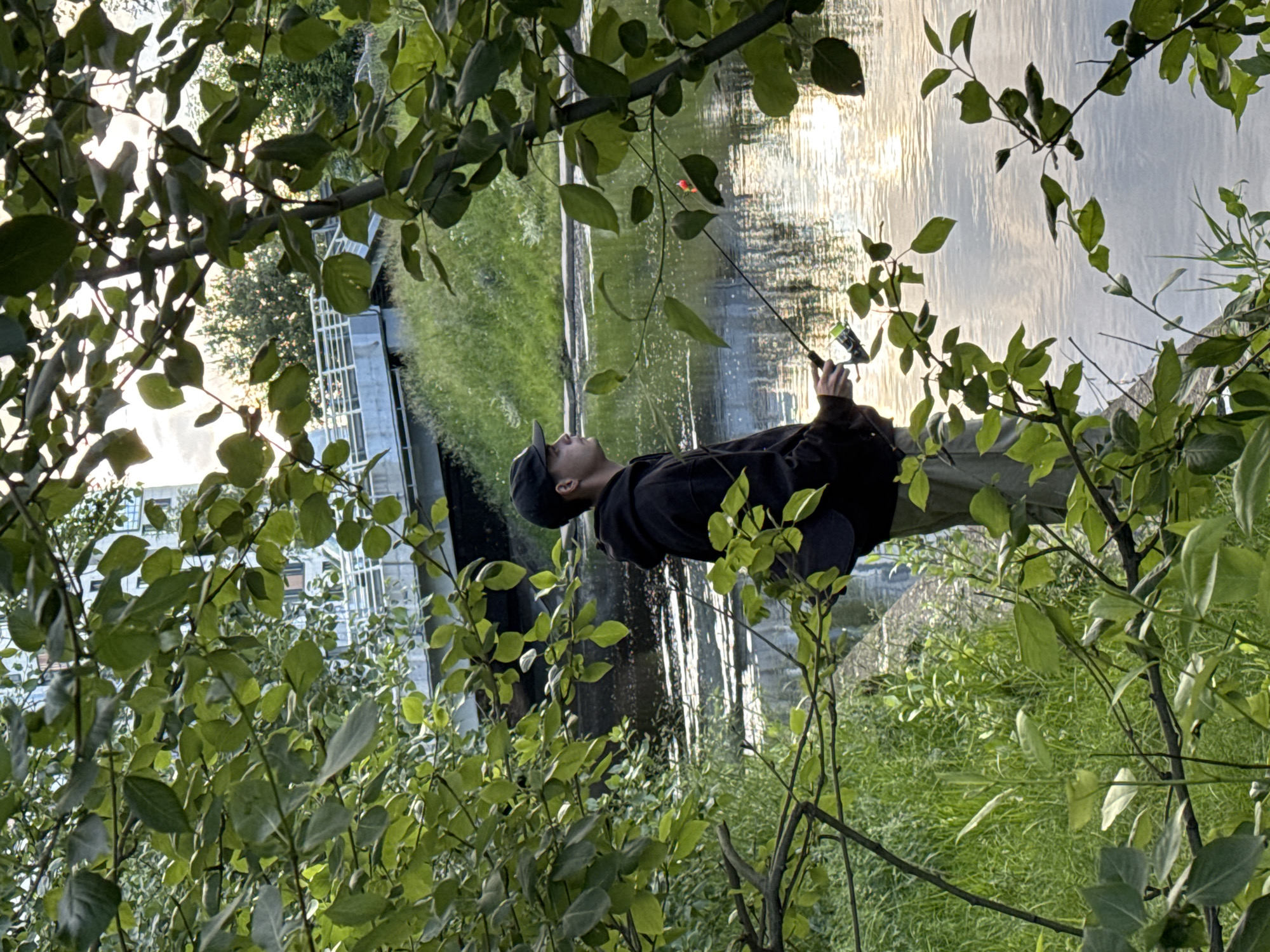A young male wearing a black baseball cap, a black hooded sweatshirt, and tan pants is fishing by a body of water. He is holding a fishing rod and looking out at the water. The water reflects the sky and sunlight, creating a shimmering effect. The background shows lush green grass, trees, and a bridge with a building in the distance. The location is believed to be Saint Petersburg, Russia, possibly near a park or a waterfront area. The overall scene depicts a serene moment of recreational activity in a natural setting within an urban environment.