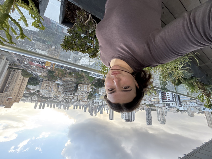A female individual is depicted in a selfie, positioned on an elevated outdoor balcony or terrace in Bnei Brak, Israel. She has dark hair and wears a dark brown long-sleeved top. Her face is directed towards the camera, with her lips pursed in an expressive manner; a thin, dark strand of material or hair is horizontally across her mouth area. The foreground features a glass railing with a metallic top bar, behind which several potted plants, including succulents, are arranged. The background reveals a dense urban cityscape, characteristic of Bnei Brak, with a broad view extending towards the skyline of Tel Aviv, identifiable by numerous modern high-rise buildings and skyscrapers in the distance. Mid-ground shows various multi-story residential and commercial buildings, some appearing to be under construction, alongside streets with visible vehicular traffic. The sky is partially overcast with grey clouds, but areas of sunlight are visible towards the horizon, suggesting late afternoon or early evening.