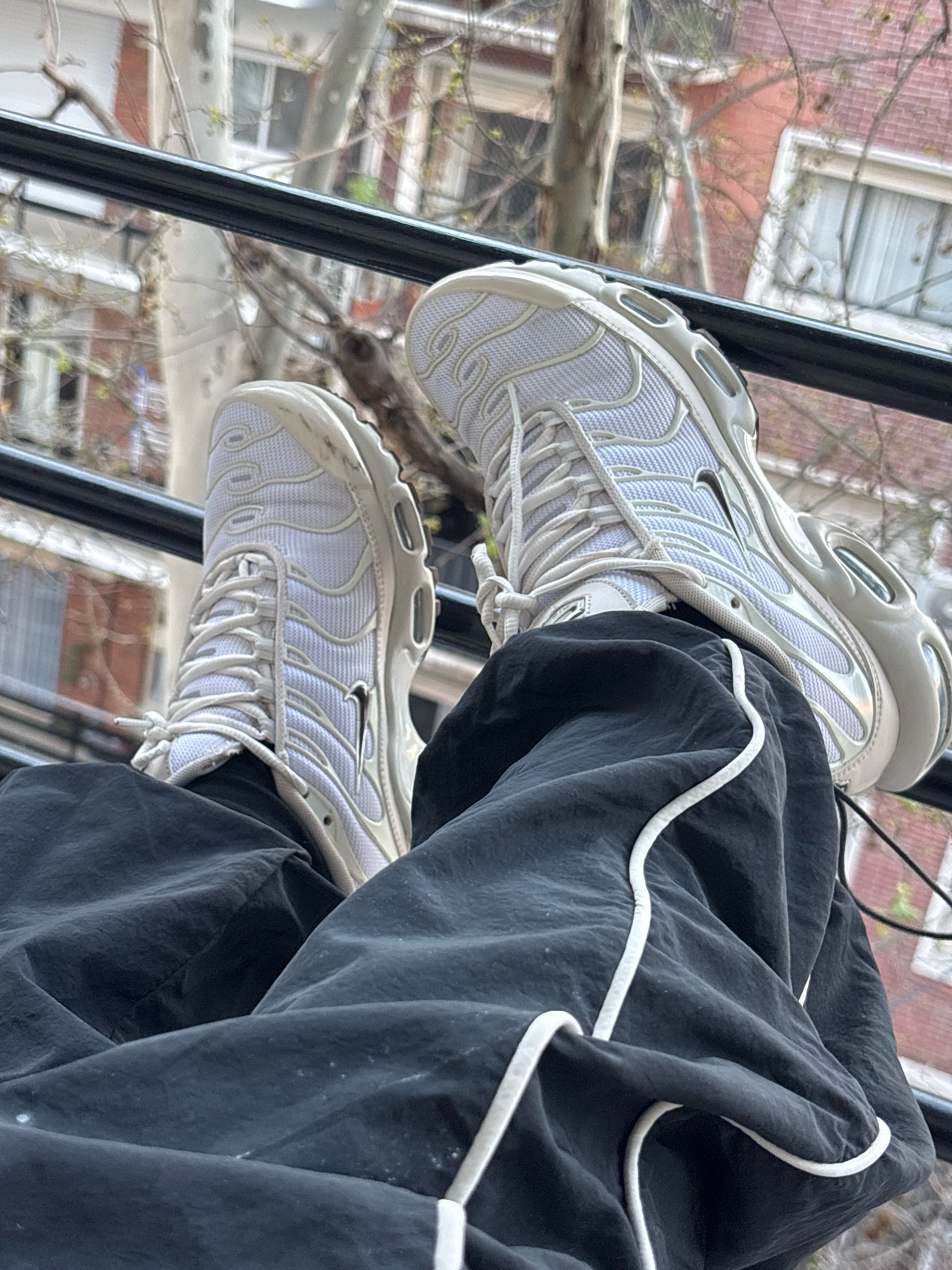 The image shows a close-up of a person's legs wearing black pants and white athletic shoes. The shoes appear to be Nike Air Max Plus TN sneakers, with a white mesh upper and light grey accents. The pants are dark black and have white piping along the seams. The person is sitting, with their legs propped up on a black railing. In the background, a building with red brick and white-framed windows is visible, along with trees that appear to be bare or have new spring foliage. The overall scene suggests an urban outdoor setting, possibly a balcony or a windowsill overlooking a street or courtyard. The location is not identifiable as Buenos Aires, Argentina.