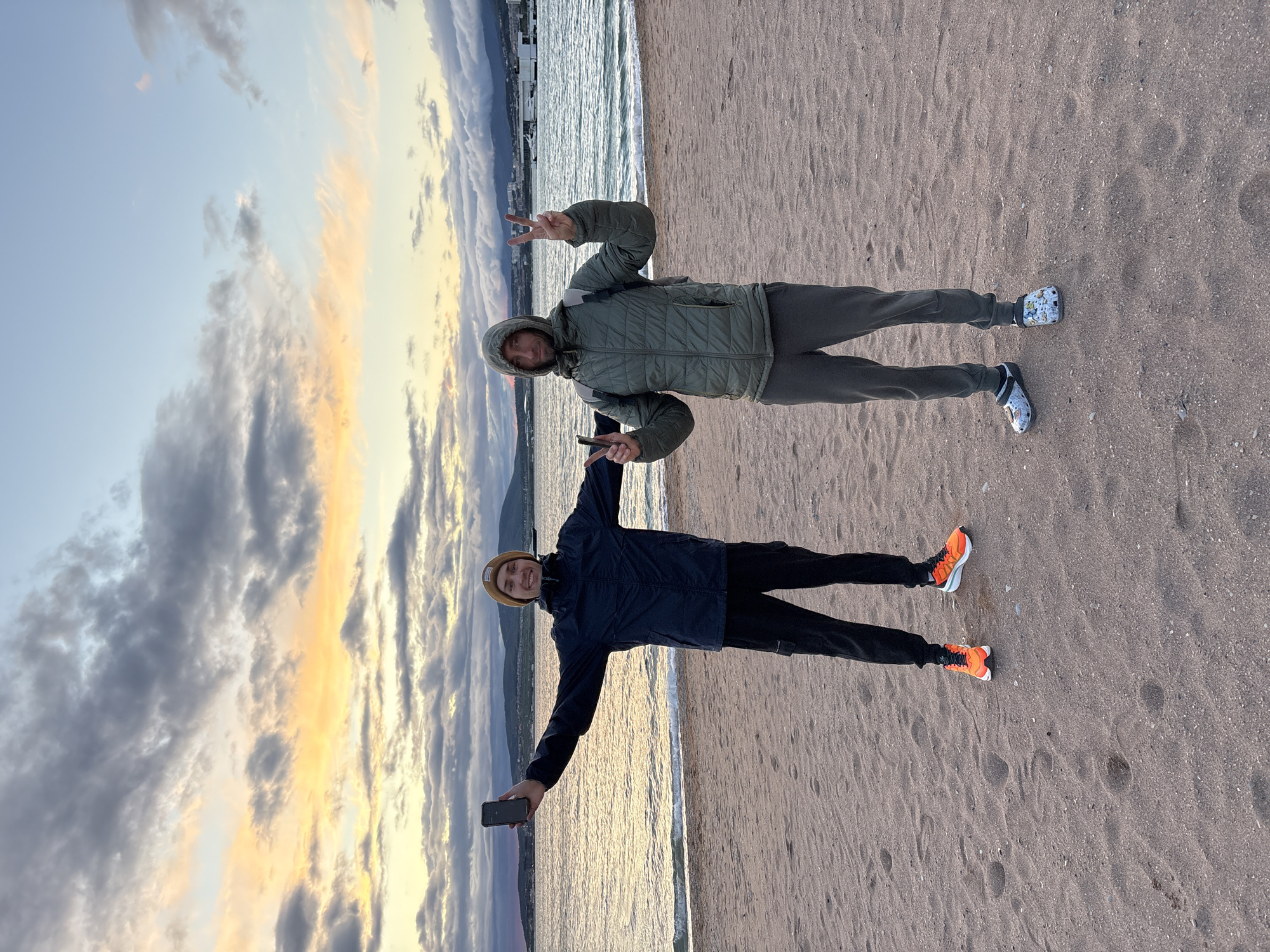 Two men are posing on a sandy beach. The man on the top is wearing a green hooded puffer jacket, grey sweatpants, and patterned Crocs. He is holding up a peace sign with both hands. The man on the bottom is wearing a navy blue jacket, black pants, a tan beanie, and orange sneakers. He is holding a black smartphone in his left hand with his arm extended. The background shows the sea, clouds, and the coastline. The location is likely Gelendzhik, Russia.