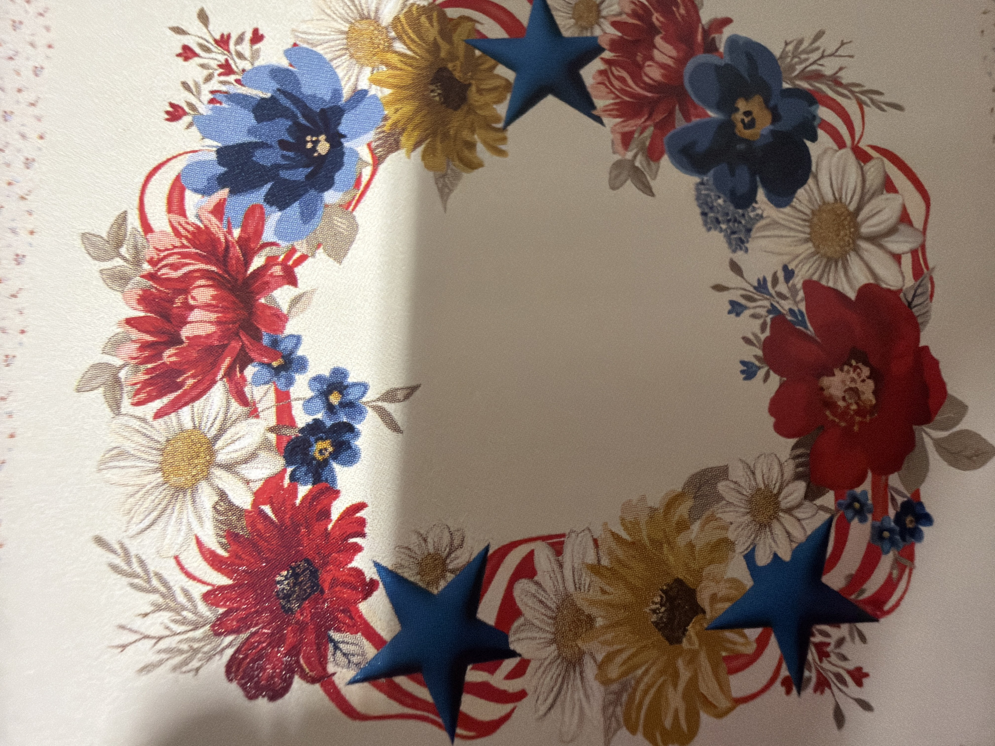 This is a close-up, overhead shot of a decorative circular wreath on a light-colored, textured fabric surface. The wreath is composed of a variety of artificial flowers in red, white, blue, and yellow, interspersed with green leaves and small sprigs. Some of the flowers are large and bold, like dahlias or chrysanthemums, while others are smaller and more delicate, resembling daisies or forget-me-nots. Intertwined with the floral elements are red and white striped ribbon-like accents, reminiscent of the American flag. Scattered throughout the wreath are three-dimensional blue stars, adding a patriotic touch. The fabric background has a subtle woven pattern and a border of very small, light pink floral-like motifs on the left side. The lighting suggests an indoor setting, possibly a table runner or decorative cloth, with a shadow cast from the top left. There is no visible text. The overall theme appears to be patriotic, suitable for holidays like the Fourth of July or Memorial Day.