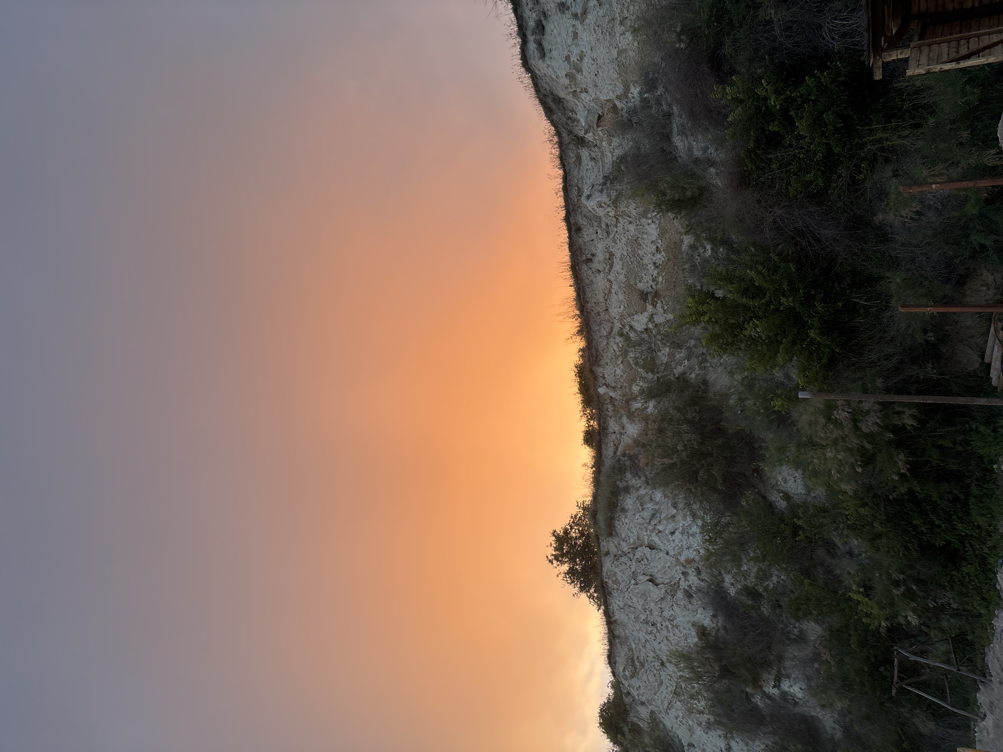 The image depicts a sunset over a cliff face covered in vegetation in Topola, Bulgaria. The sky displays a gradient of colors, transitioning from a soft lavender at the top to a vibrant orange near the horizon. The cliff itself is composed of light-colored rock and is partially covered with dry grass and scattered green bushes and trees. At the bottom of the image, a wooden structure, possibly a small shed or lean-to, is visible on the right side. On the left, part of a rudimentary swing set can be seen, along with some wooden logs and fencing. The scene appears to be an outdoor, natural environment at dusk.