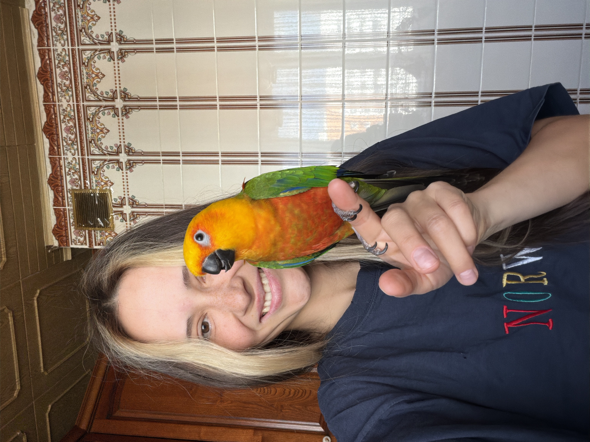 A young woman with blonde streaks in her dark hair and freckles across her nose smiles at the camera while holding a bright orange and green parrot on her finger. The parrot is perched on her left index finger, its claws visible against her skin. The woman is wearing a dark blue t-shirt with embroidered text that appears to say "NORWAY" in colorful letters. She is standing indoors, with a wooden door or cabinet visible to her left and a tiled wall with an ornate pattern behind her. The ceiling above is paneled wood. The overall scene appears to be domestic, possibly a home environment. The image does not provide any specific information or clues about the city of Khimki, Russia.