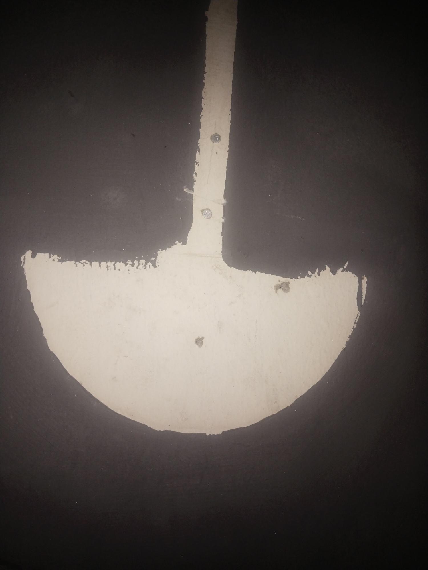 This is an eye-level indoor shot of a black wall featuring a white, shovel-shaped figure. The shovel consists of a vertical handle and a semi-circular spade. Three screws are visible along the handle's length. The paint application on the spade is uneven, with rough edges suggesting a partially removed or poorly applied coat. The black wall shows texture, and small blemishes can be seen across both the painted and unpainted surfaces. No people or other objects are present. The location is stated as Unknown, Unknown.