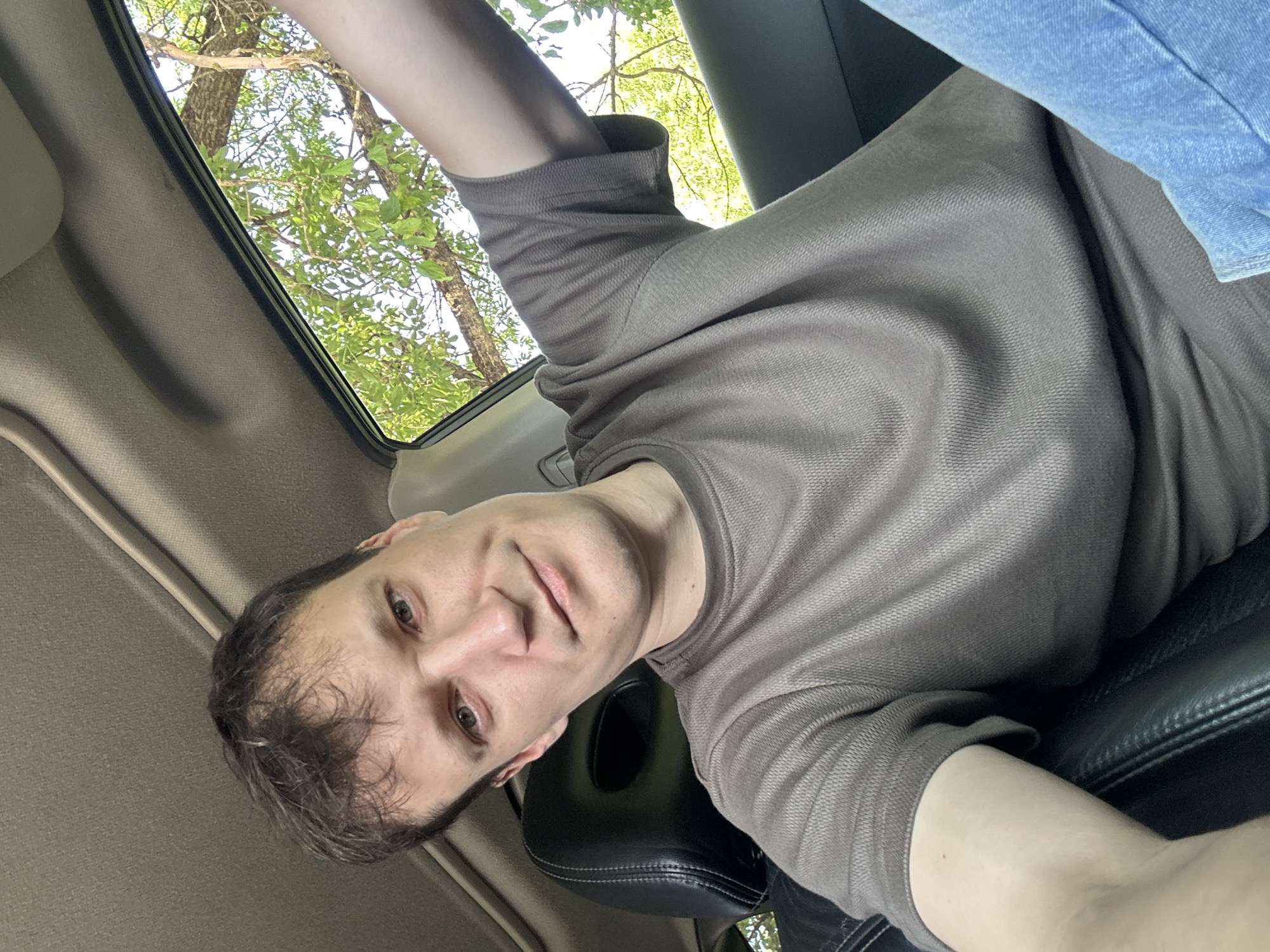 A man with light skin and short, dark brown hair is taking a selfie in a car. He is wearing a brown t-shirt and blue jeans. The interior of the car is visible, including a black leather seat and the car's headliner. Sunlight streams in from a window, illuminating the man's face and the green leaves of a tree outside. The man's left arm is extended, likely holding the camera. The scene appears to be during the daytime. The location is Makiv, Ukraine.