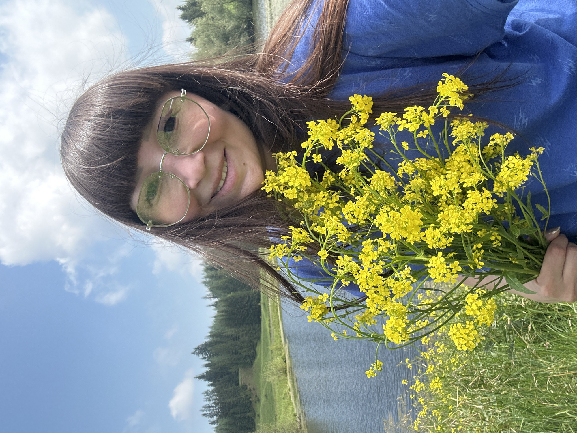 In Buranovo, Russia, a woman with long, dark hair and bangs, wearing round, light-tinted glasses and a blue t-shirt, holds a bouquet of yellow wildflowers. She is smiling and looking at the camera. The background shows a tranquil lake, lush green grass, and a dense forest of evergreen trees under a partly cloudy blue sky. The overall scene evokes a sense of natural beauty and peacefulness.