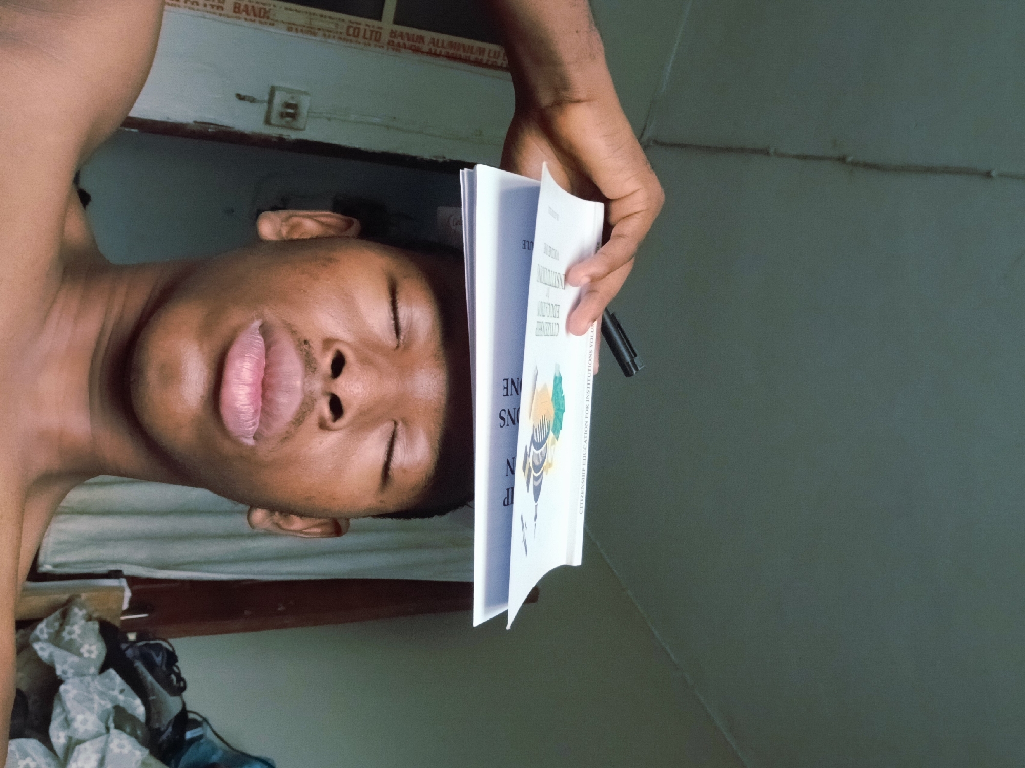 A shirtless man is holding a book titled "Citizenship Education for Institutions" near his face and a black pen is clipped to the edge of the book. The OCR text includes "CO LTD BANUR ALLAMINIUM LU" and "BAND DA" which potentially indicates the location in the city of Lagos, Nigeria.