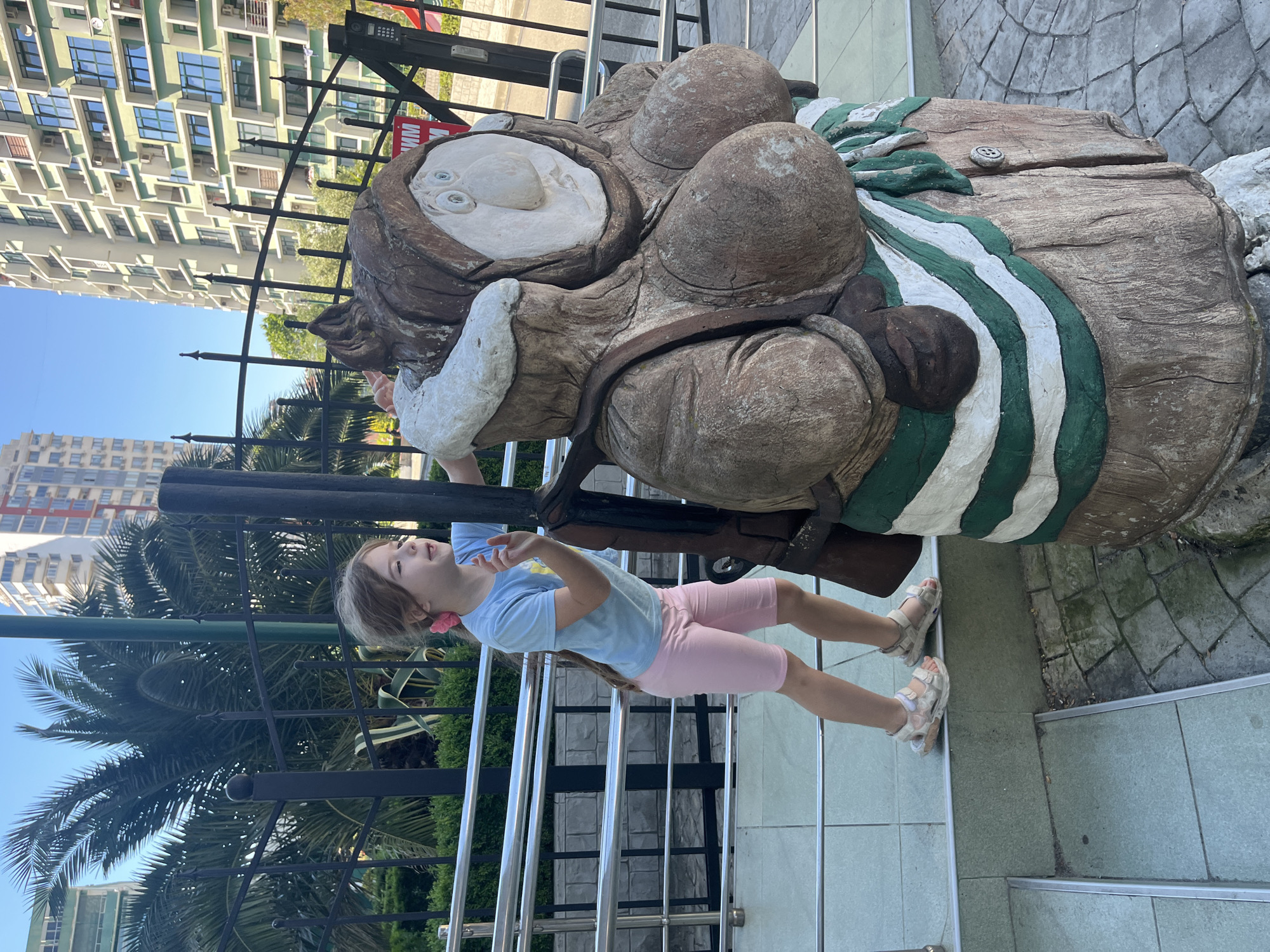A young Caucasian girl with blonde hair tied back with a pink bow, wearing a light blue t-shirt and pink shorts, stands on a tiled step looking at a large, wooden sculpture of a woman. The sculpture has a brown, textured surface and appears to be wearing a hooded garment with green and white stripes around its waist. The girl's left hand is extended towards the sculpture, and her right arm is bent upwards. Behind the girl and sculpture, there is a black metal fence, palm trees, and modern, multi-story buildings with light-colored facades and green-tinted windows. The ground in front of the sculpture is paved with light-colored interlocking stones. The scene appears to be outdoors, likely in a public area within the city of Sochi, Russia, given the architectural style of the buildings and the presence of palm trees. The overall lighting suggests it is daytime, with clear skies.