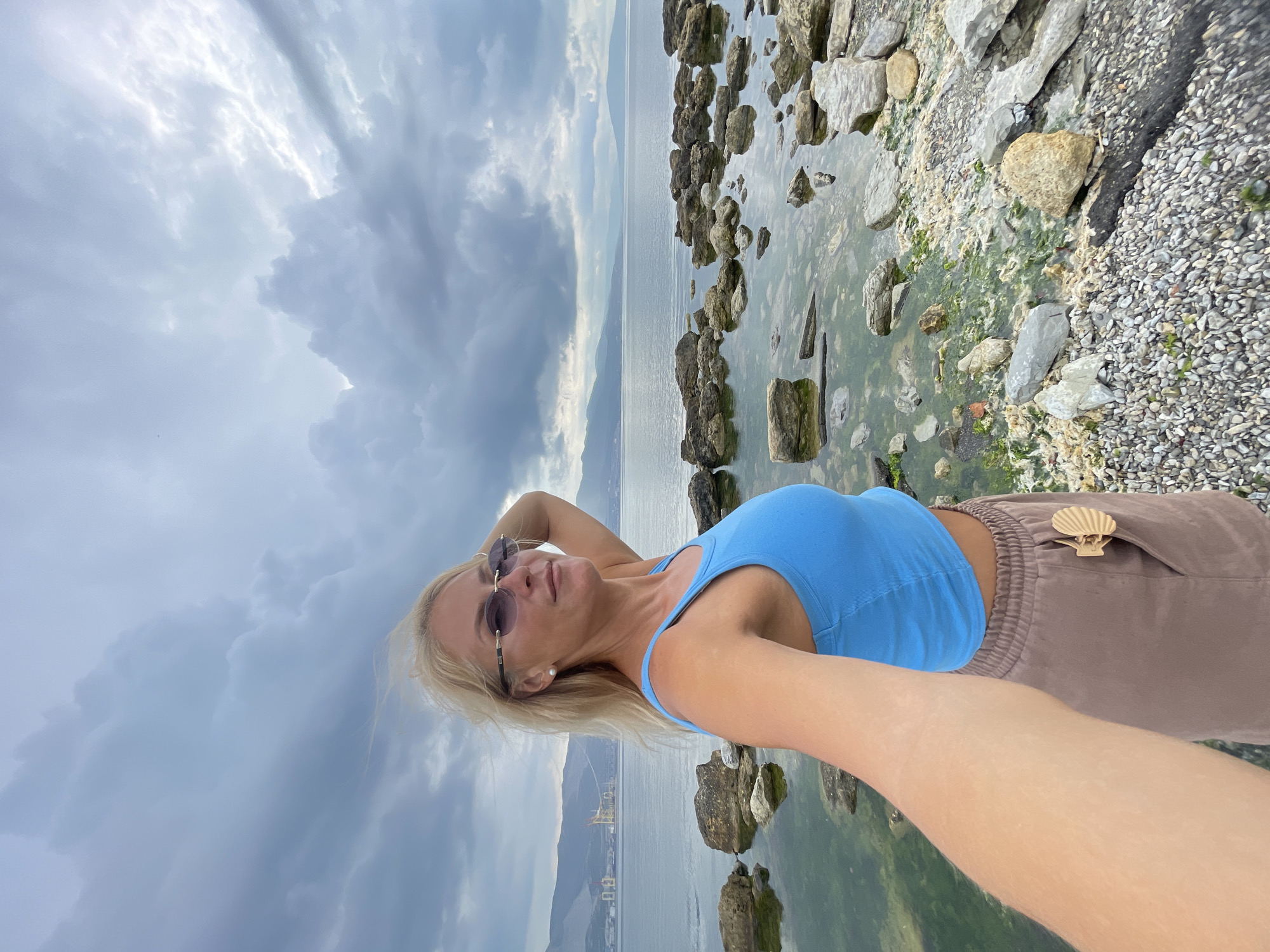 A blonde woman with sunglasses takes a selfie on a rocky shoreline. She is wearing a blue tank top and brown shorts. The sea is calm and the sky is cloudy. In the background, there are mountains and industrial structures. The location is the coast of Novorossiysk, Russia.