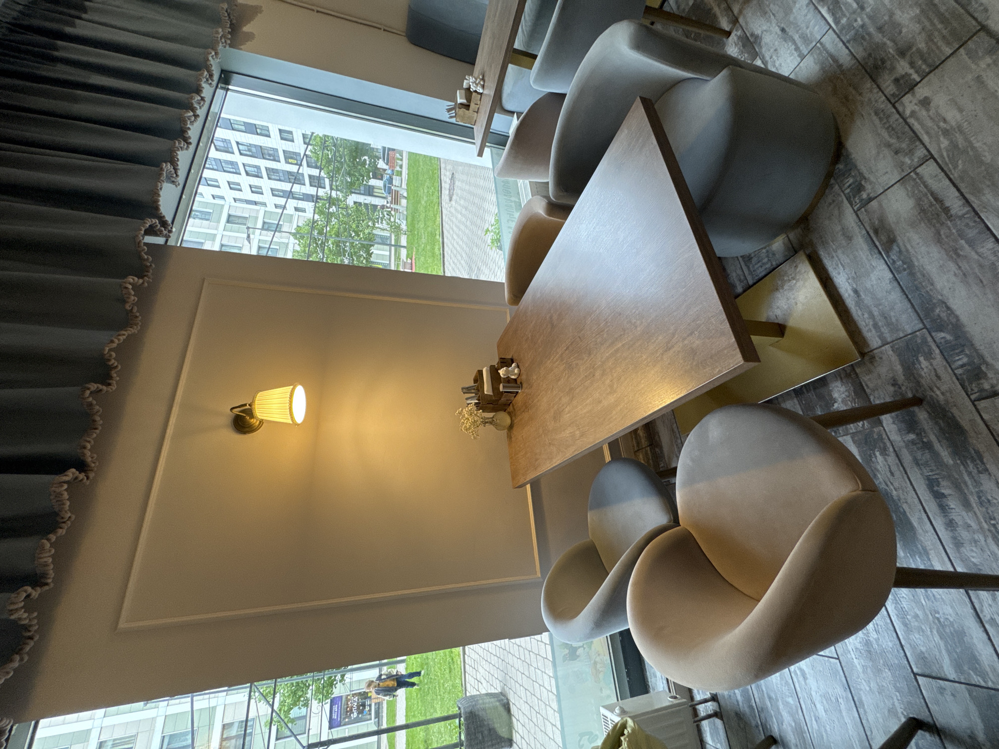 The image captures an interior scene of what appears to be a cafe or restaurant setting. The viewpoint is from above, looking down onto a table arrangement.
The primary focus is a square, wooden table, which is part of a set. Several chairs with a modern design are positioned around the table. The chairs are upholstered, with a curved back and armrest design.
The environment shows a modern, stylish interior. A window provides a view of a cityscape, which includes apartment buildings and trees. This gives the impression that the location is in a relatively urban area. A lamp is mounted on the wall.
Based on the urban environment visible through the window, it's reasonable to hypothesize that the image was taken in Saint Petersburg, Russia.