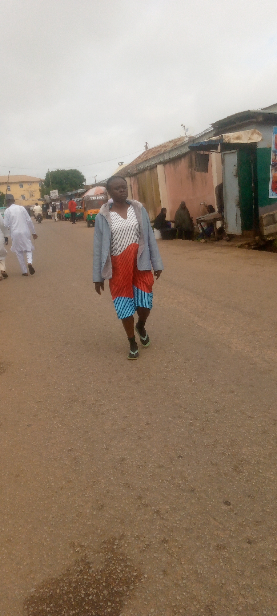 A woman walks down a dusty street, wearing a light blue jacket over a striped and orange-patterned dress, and black flip-flops. She is in the foreground, facing slightly to the right. In the background, several people are visible, some walking and others sitting near buildings. A yellow auto-rickshaw is also present on the street. The environment appears to be an urban setting with buildings lining one side of the road and an overcast sky overhead.