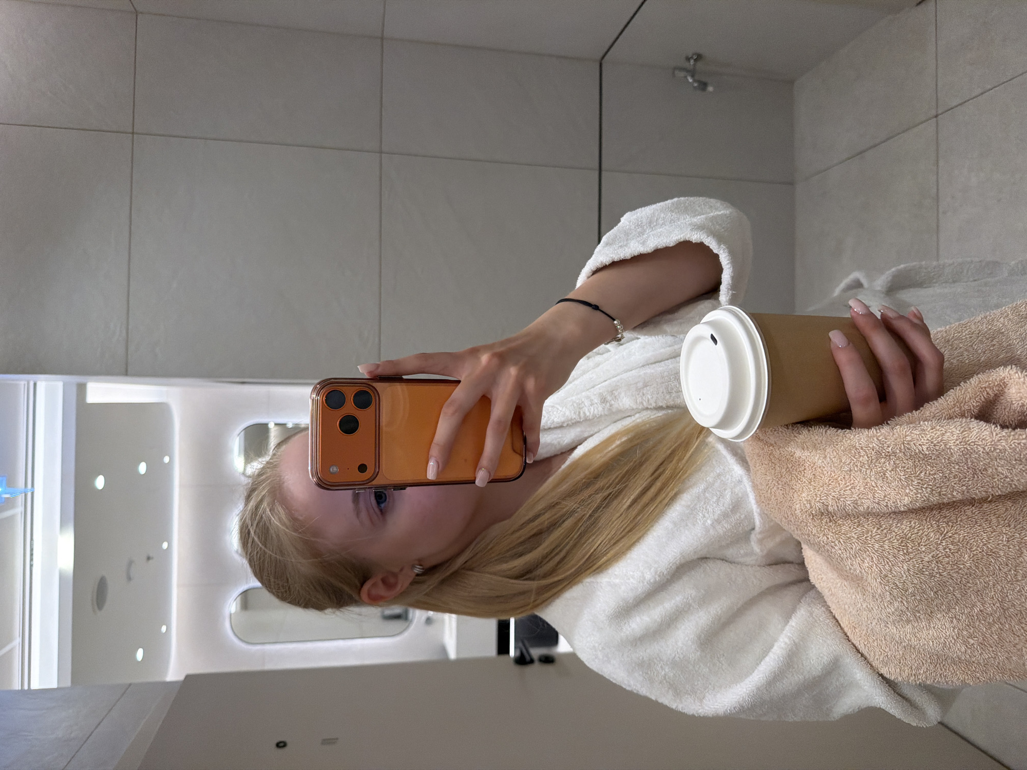 A person with long blonde hair, wearing a white bathrobe, is taking a selfie in a bathroom. The person is holding an orange smartphone in one hand and a brown disposable coffee cup with a white lid in the other hand. The bathroom features white tiles, a mirror, and a shower cabin with multiple lights. The location appears to be indoors.