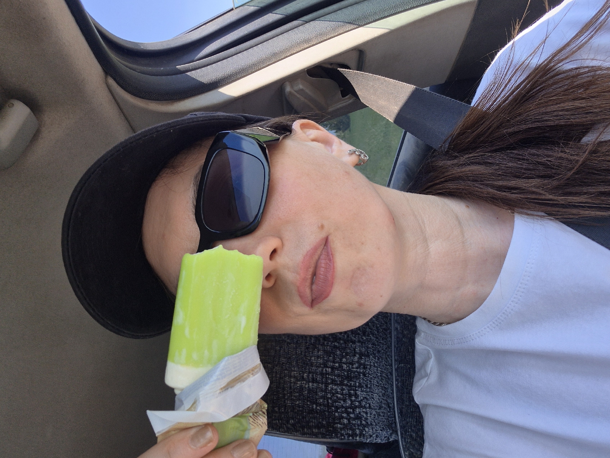 The image shows a woman wearing a black hat and sunglasses while inside a vehicle. She has a seatbelt fastened across her body and is holding a green popsicle with a white bottom portion, partially unwrapped. Her hair is dark brown and partially covers her shoulder. The interior of the car is visible, including part of the roof and dashboard. The location in the city is Unknown, Unknown.