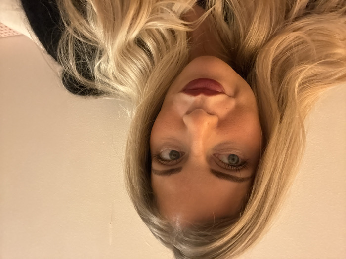 A woman with blonde, wavy hair poses close to the camera. Her eyes are light-colored, and she wears dark lipstick. She appears to be fair-skinned and is looking directly at the camera. The background is a neutral, light color. The photo angle is slightly tilted. A dark garment, possibly a top, is visible at the top of the image.