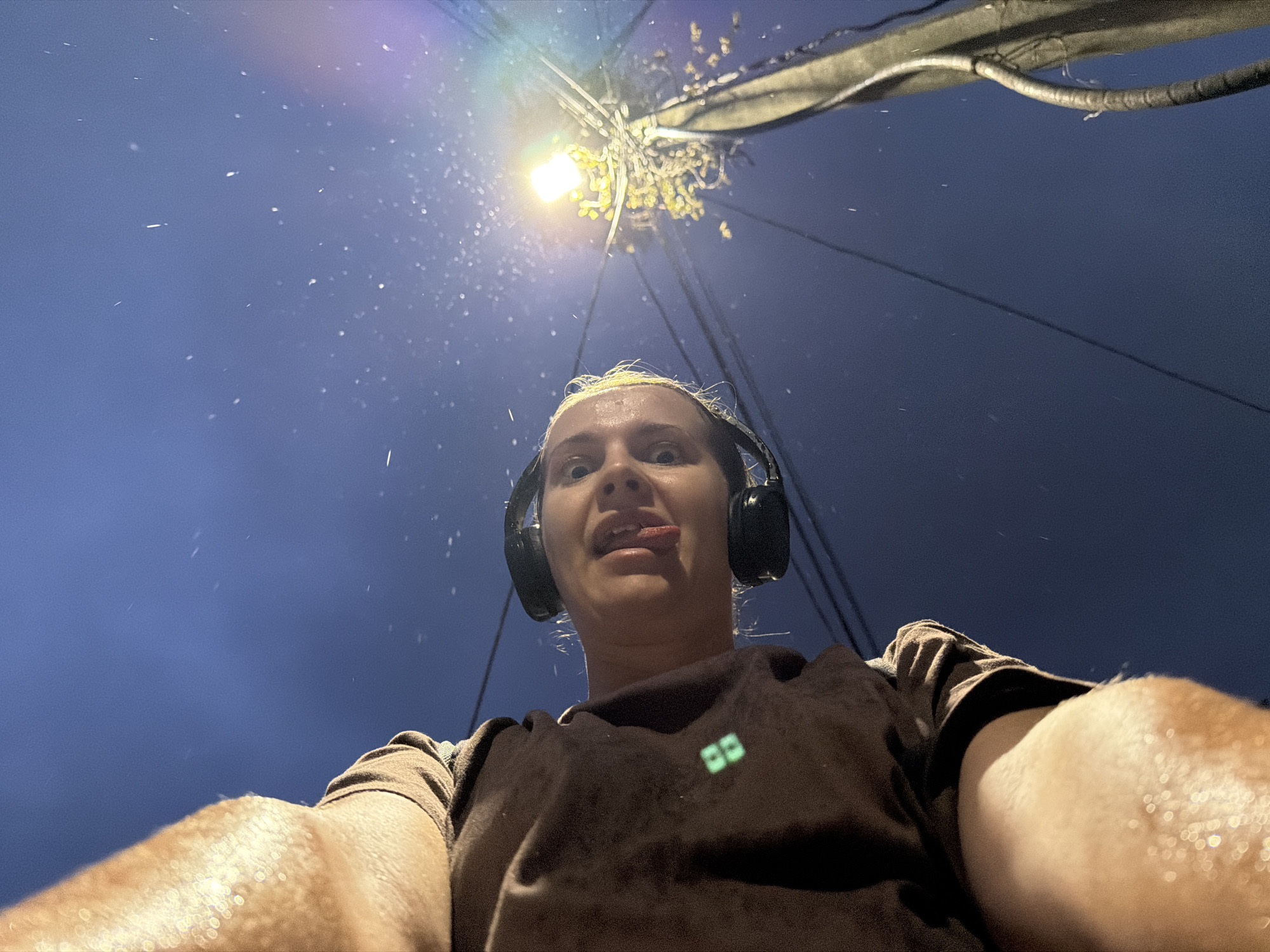 In a selfie taken from a low angle outdoors at night, a person with blonde hair wears black headphones and a brown sweatshirt with a small green logo on the left chest. The person is sticking their tongue out, looking towards the camera. The background is a dark blue sky with many small white specks, possibly snow or rain, illuminated by a bright yellow street light on a utility pole. The pole has black wires attached to it, and some foliage is visible around the light. The person's arms and shoulder are visible in the foreground, appearing wet or sweaty. The scene is consistent with a location in Sochi, Russia, during a winter evening with precipitation and street lighting.