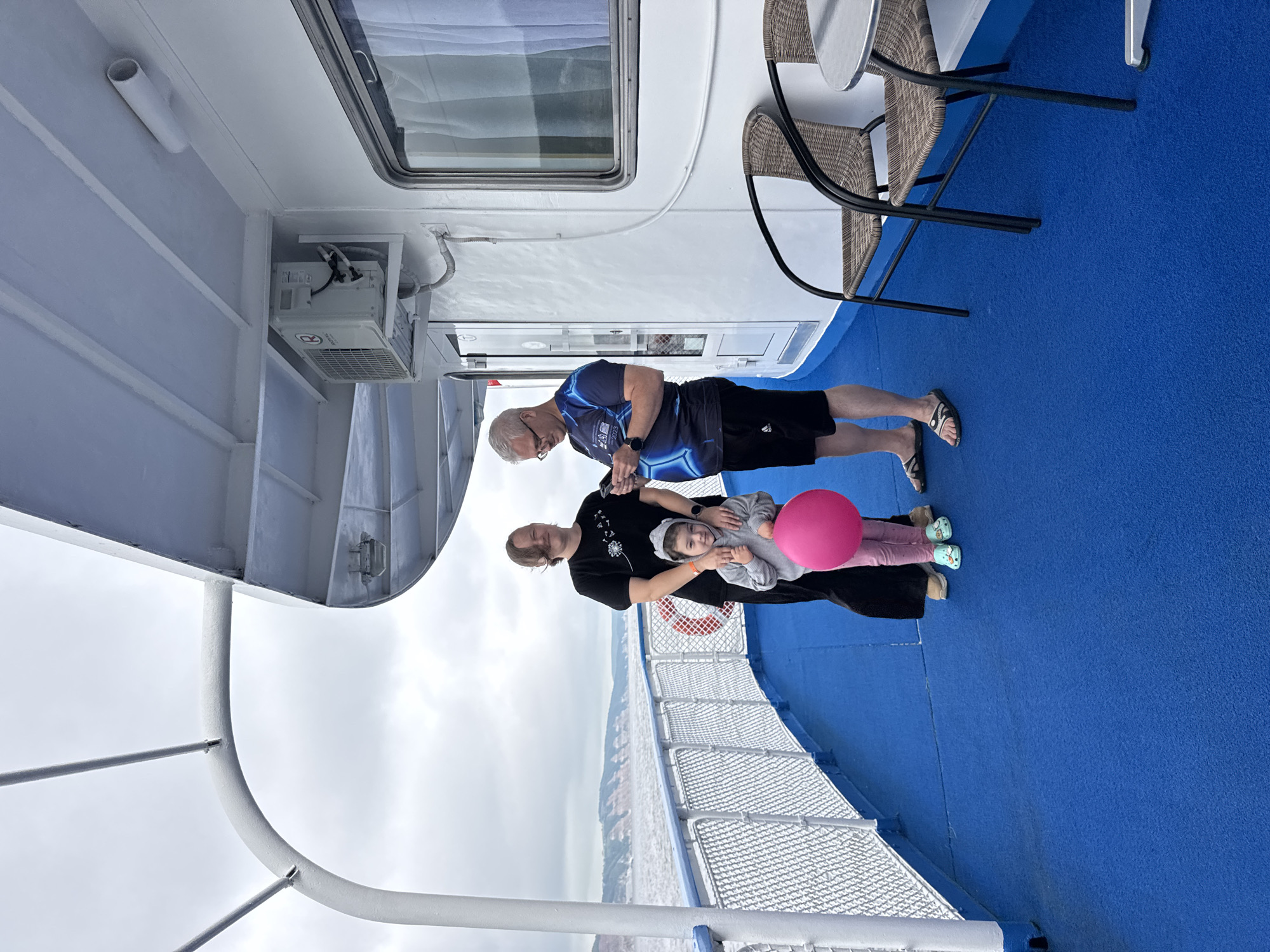 A family is posing for a photo on the deck of a boat, likely in Kamskoye Ustye, Russia, given the presence of the Volga River and the distinctive cliff formations in the background. A woman on the left, dressed in a black t-shirt and dark pants, stands with her arm around a young girl. The girl, wearing a gray hooded sweatshirt and pink leggings, holds a bright pink balloon and looks at the camera. To the right, a man in a blue and black t-shirt and black shorts, wearing glasses, is looking down at his phone. The deck is covered in a vibrant blue carpet, and a white railing runs along the edge, with the water and distant cliffs visible beyond. Two wicker chairs and a small table are also present on the deck, suggesting a leisure setting. An air conditioning unit is mounted on the white exterior wall of the boat. The sky is overcast.