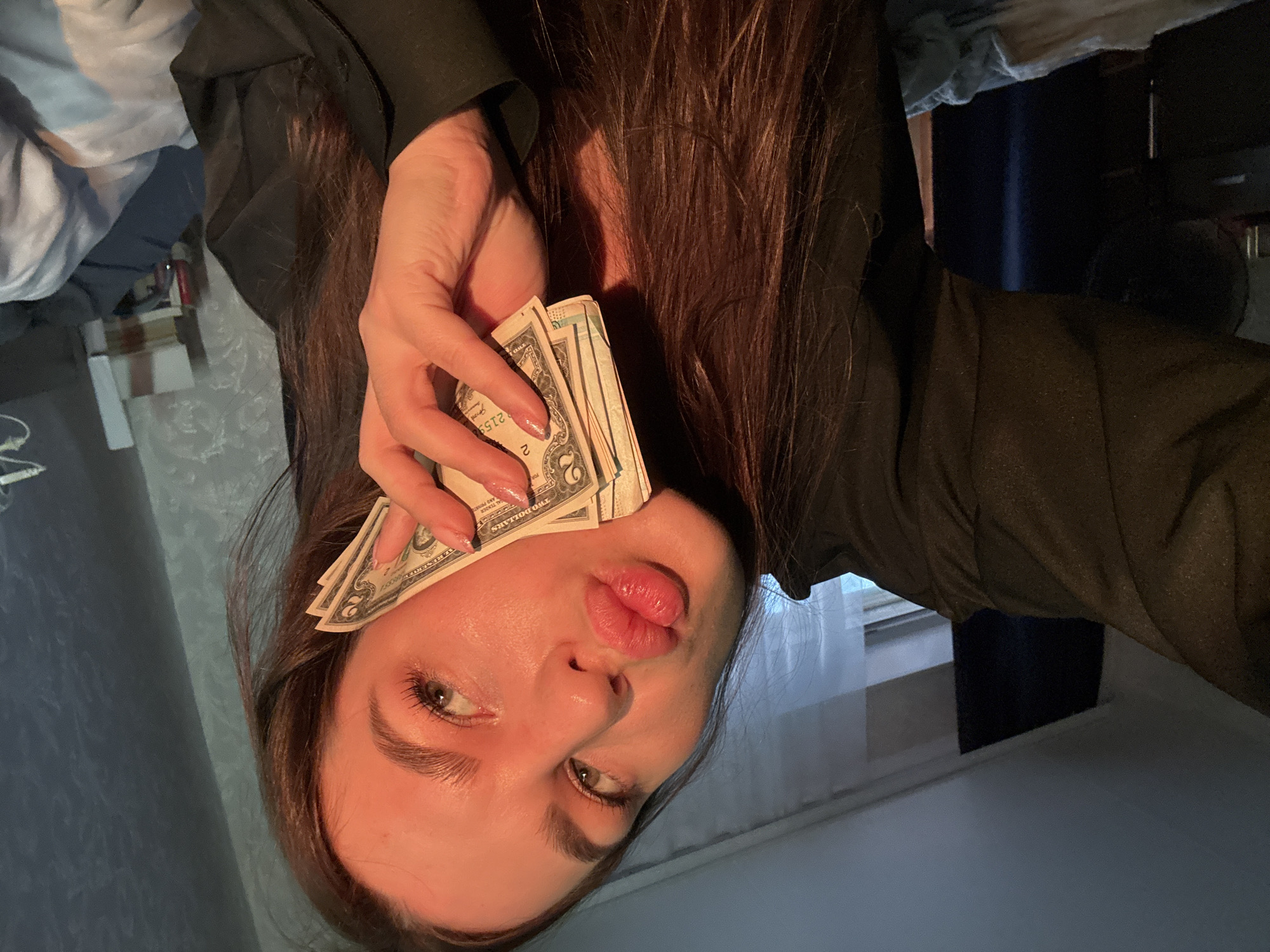A young woman is taking a selfie in a dimly lit room. She is holding a fan of dollar bills to her cheek, and her lips are pursed in a kiss. The room appears to be a bedroom, with a bed and curtains visible in the background. The lighting is warm and orange, suggesting it may be evening or the room is lit by artificial light. The woman has long, dark hair and is wearing a dark top. The scene does not provide any specific indicators that place it within the city of Chaikovsky, Russia.