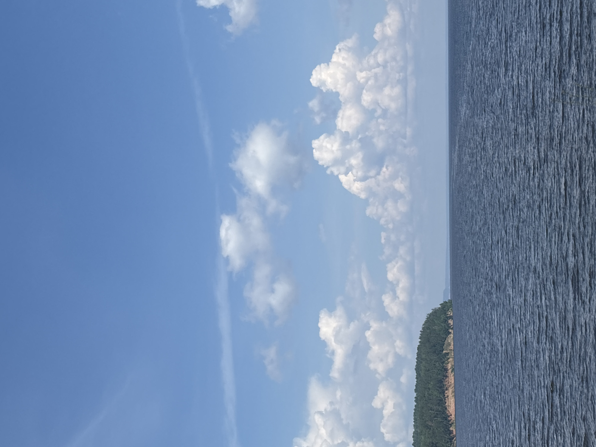 The image depicts a wide expanse of dark blue water under a bright blue sky dotted with fluffy white clouds. On the left side of the frame, a lush green forest covers a steep, reddish-brown embankment that juts out into the water. The water's surface is textured with ripples. The scene captures a serene natural landscape. This appears to be a view of the Kama River near the city of Chaikovsky, Russia, a region known for its expansive waterways and forested shores. There are no people or discernible man-made structures visible in the frame.
