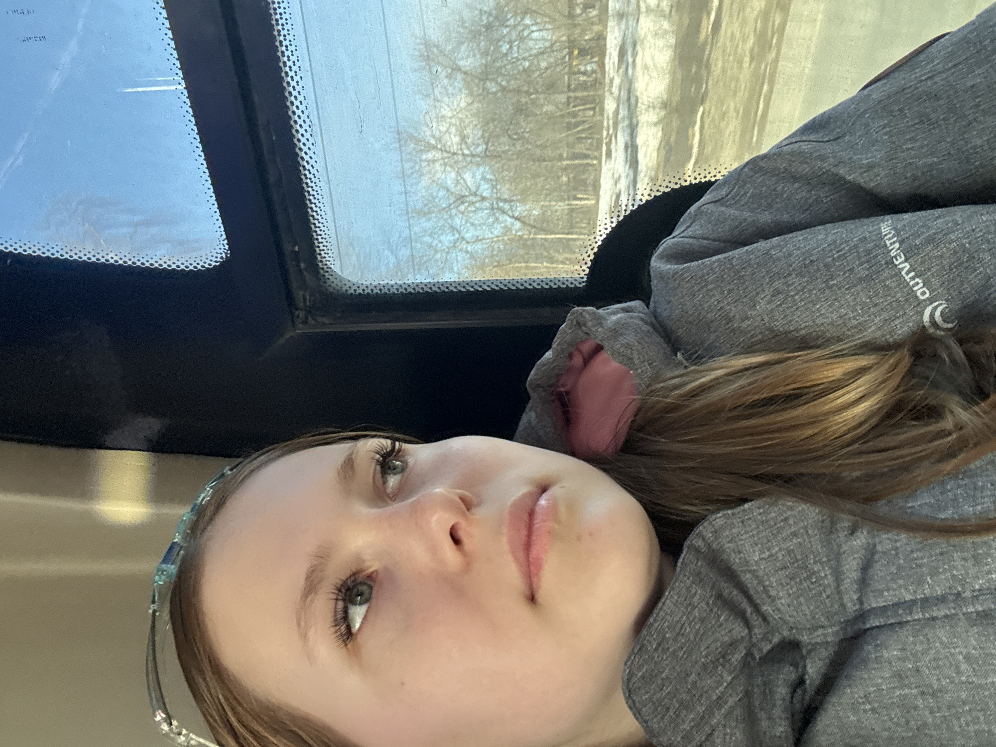A close-up shot depicts a young woman with light skin and green eyes, framed by light brown hair. She wears a headband and a gray hooded jacket with the word "OUTVENTURE" printed on the sleeve. The background shows an interior view with a window, revealing leafless trees and a snowy landscape. The location is likely Berezniki, Russia, as inferred from the potential metadata. The scene evokes a sense of stillness, as the woman appears contemplative while looking out the window.