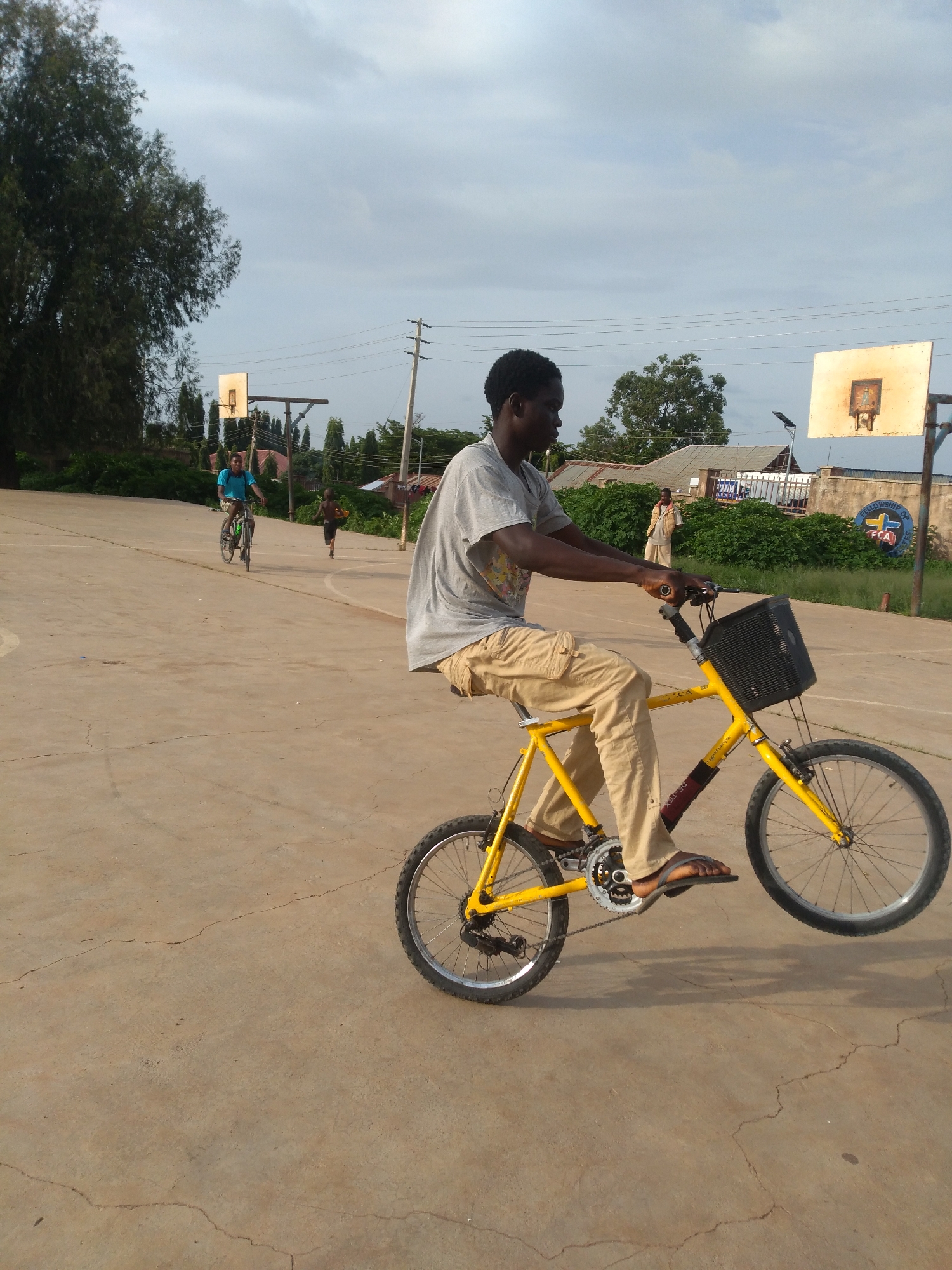 A young Black man, wearing a gray t-shirt and tan cargo pants, is doing a wheelie on a yellow bicycle on a concrete basketball court. The bicycle has a black basket on the front. The man is wearing sandals on his feet. In the background, another young Black boy in a blue shirt is riding a bicycle, and a younger Black boy is running with a basketball. Two adults are standing in the background near a building. There are basketball hoops and power lines in the distance. The sky is cloudy.
