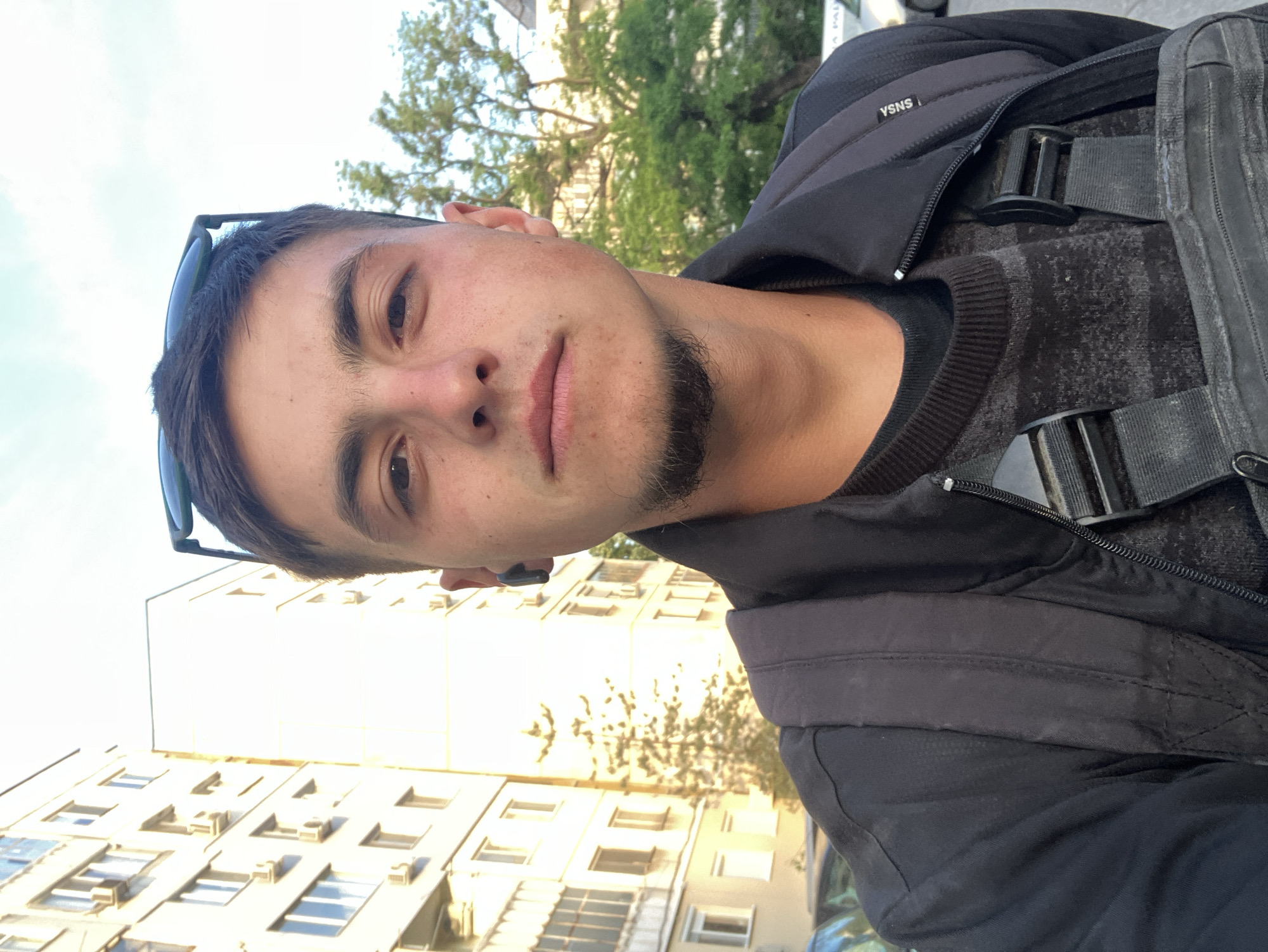 A young man is facing the camera outdoors. He has short dark hair, stubble, and sunglasses propped on his head. He is wearing a dark jacket over a dark patterned sweater and a black backpack. Behind him, a multi-story building with many windows is visible, along with some trees. The location is likely Plovdiv, Bulgaria.