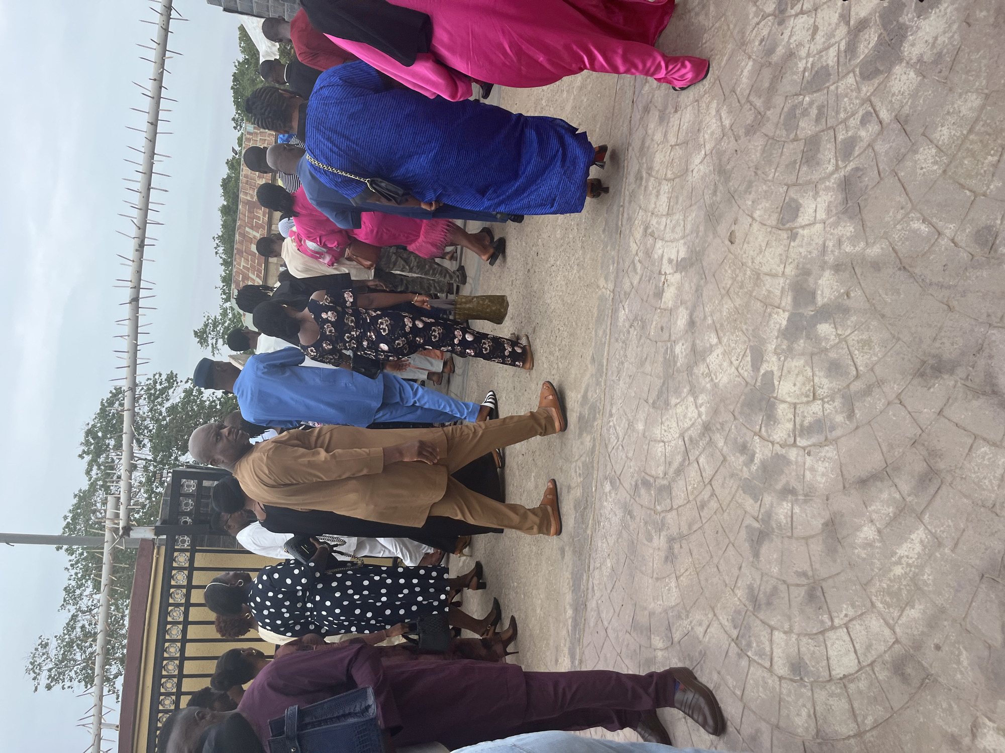 The image depicts a line of people in Abuja, Nigeria. They are diverse in attire. The environment features a gate with spikes and a paved surface. The individuals are standing, possibly waiting or queuing. Buildings and trees are visible in the background.