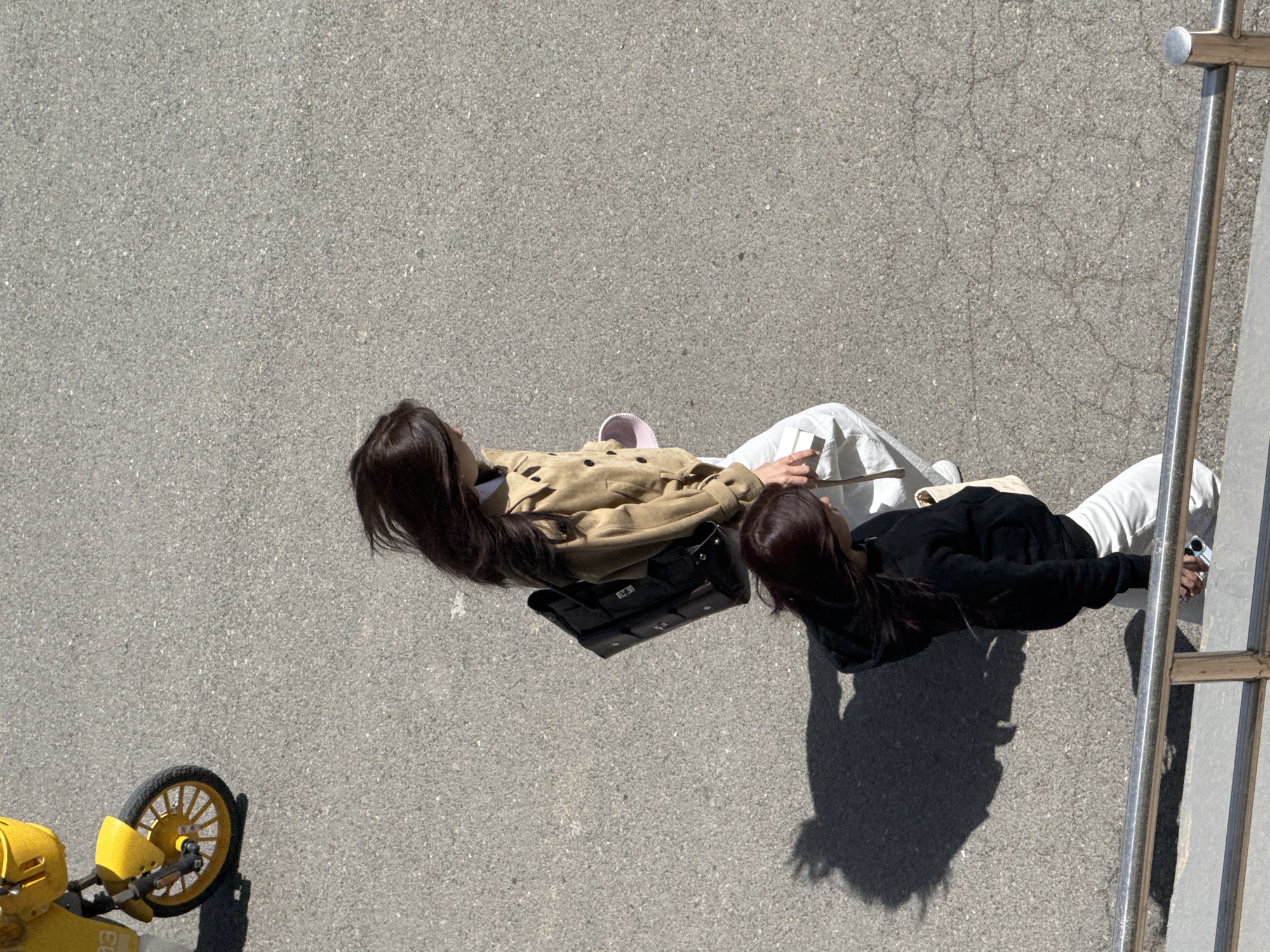 Two women are sitting on a paved surface adjacent to a metal structure. One woman is wearing a tan jacket, white pants, and carrying a black bag. The other woman is wearing a black jacket and white pants. A yellow object with a wheel is in the lower left corner. The setting appears to be outdoors in Zhongtun, China.