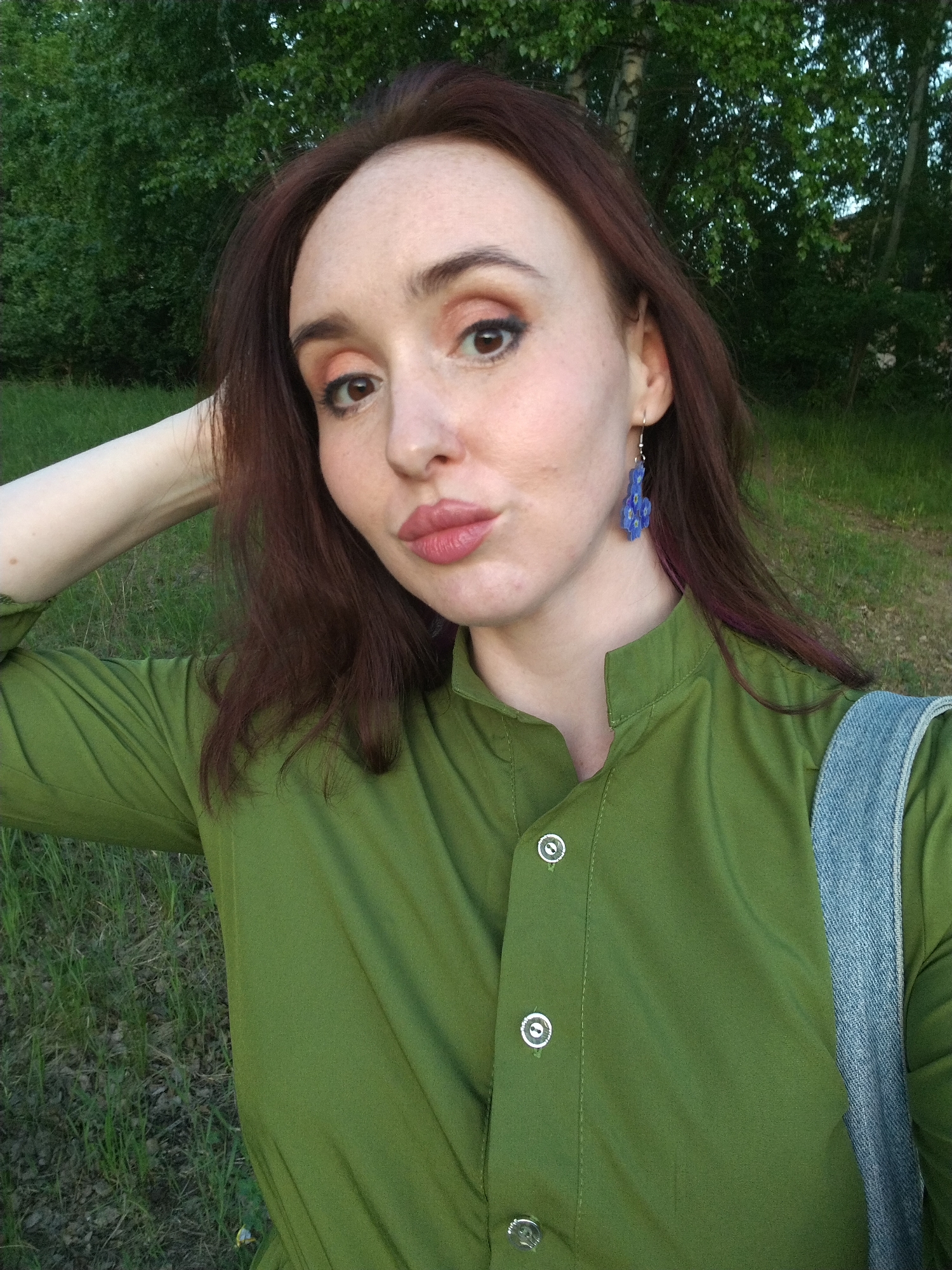 A person with reddish-brown hair and light skin poses for a selfie outdoors. The person is wearing a green collared shirt with silver buttons and blue earrings. They have one arm raised and are pursing their lips. A blue denim strap is visible on their shoulder. The background shows a grassy area with trees. The location cannot be determined as Izhevsk, Russia from the image alone.