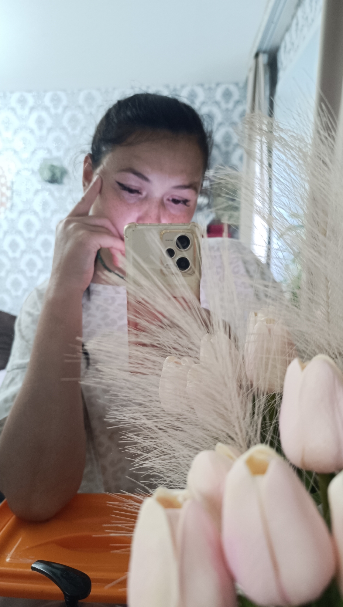 A woman with dark hair pulled back is holding a smartphone in her right hand, taking a selfie in a mirror. She is wearing a light-colored t-shirt with a subtle pattern. Her left hand is near her face, with her index finger touching her temple. She has eyeliner and light freckles on her face. In the foreground, out of focus, are several light pink tulips and wispy white decorative reeds. Behind her, the wallpaper has a patterned design. The scene appears to be indoors, possibly a home. The image quality is moderate, with some graininess. There is no indication of the specific location within the city of Chaikovsky, Russia.