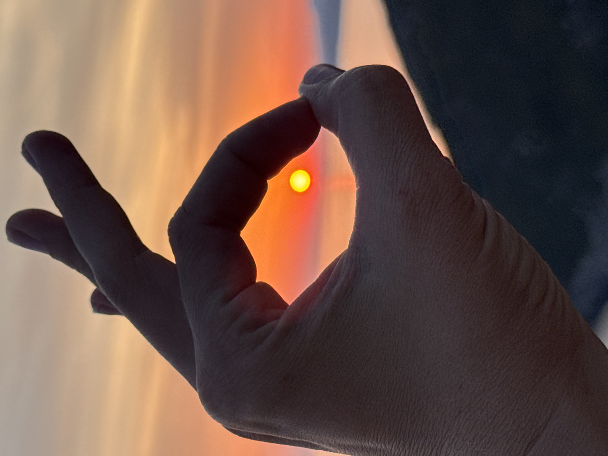 A close-up shot captures a hand forming an "OK" gesture against a vibrant sunset sky. The sun, a small, bright orb, is perfectly framed within the circle created by the thumb and index finger. The sky transitions from a warm orange near the horizon to a softer peach and yellow higher up. In the background, a hazy body of water and distant landforms are visible, silhouetted against the colorful sky. The foreground shows the textured surface of the hand, with clear details of skin creases. The dark, blurry outline of a hill or mountain slope anchors the bottom of the frame. The image is sharp and well-focused on the hand and the sun.