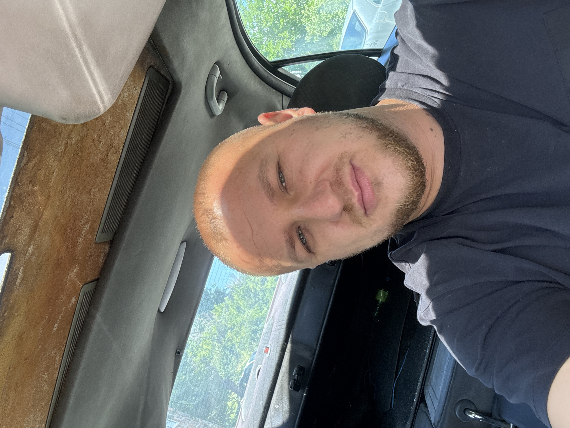 A man with a light complexion, short blonde hair, and a beard is visible in a first-person shot from inside a vehicle. He wears a dark blue t-shirt. The interior of the vehicle is visible, including part of the dashboard, the ceiling, a window, and a sun visor. Through the window, trees and parts of other vehicles are visible. The location of the photo is Unknown, Unknown.