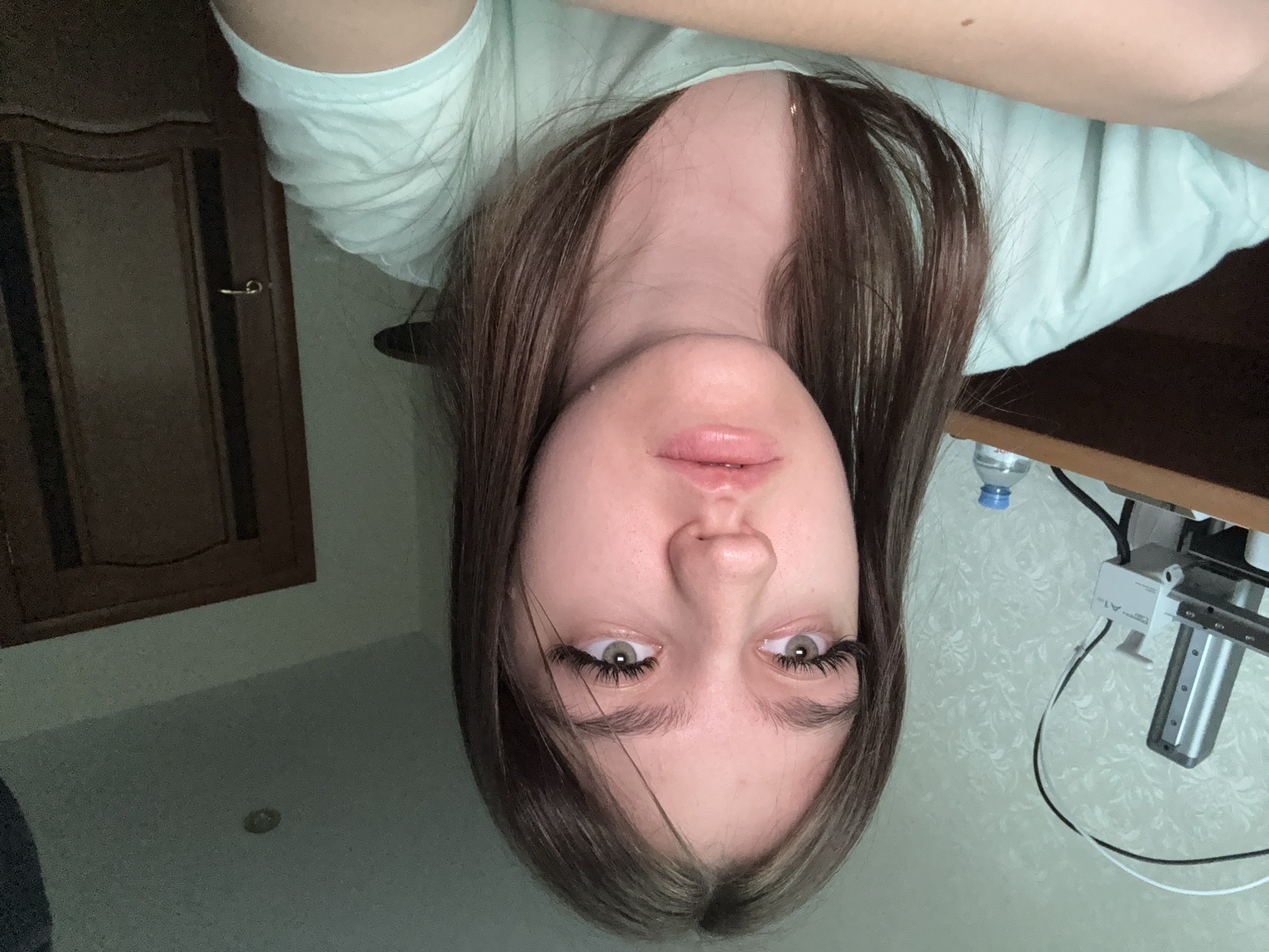 An upside-down, close-up indoor shot captures a young person, likely female, looking directly into the camera. Her head is positioned towards the bottom of the frame (when viewed correctly), with her long, straight brown hair, which has some lighter streaks, framing her face. She has green/hazel eyes with prominent, long dark eyelashes and light pink, slightly parted lips. Her expression is neutral. She is wearing a light mint green t-shirt.
In the background, on the left (when viewed correctly), a dark wooden door with a light-colored handle is visible. On the right, a light-colored wall with a subtle patterned wallpaper is seen, behind what appears to be a wooden desk or shelf. On this surface, a small clear plastic water bottle with a blue cap is present, next to a piece of silver electronic equipment with visible cables, possibly a component of a printer, scanner, or an adjustable mount. The text "A1" is faintly visible on this equipment. The overall lighting suggests an indoor setting, possibly with artificial light. The person appears to be lying down or leaning back, looking up at the camera.