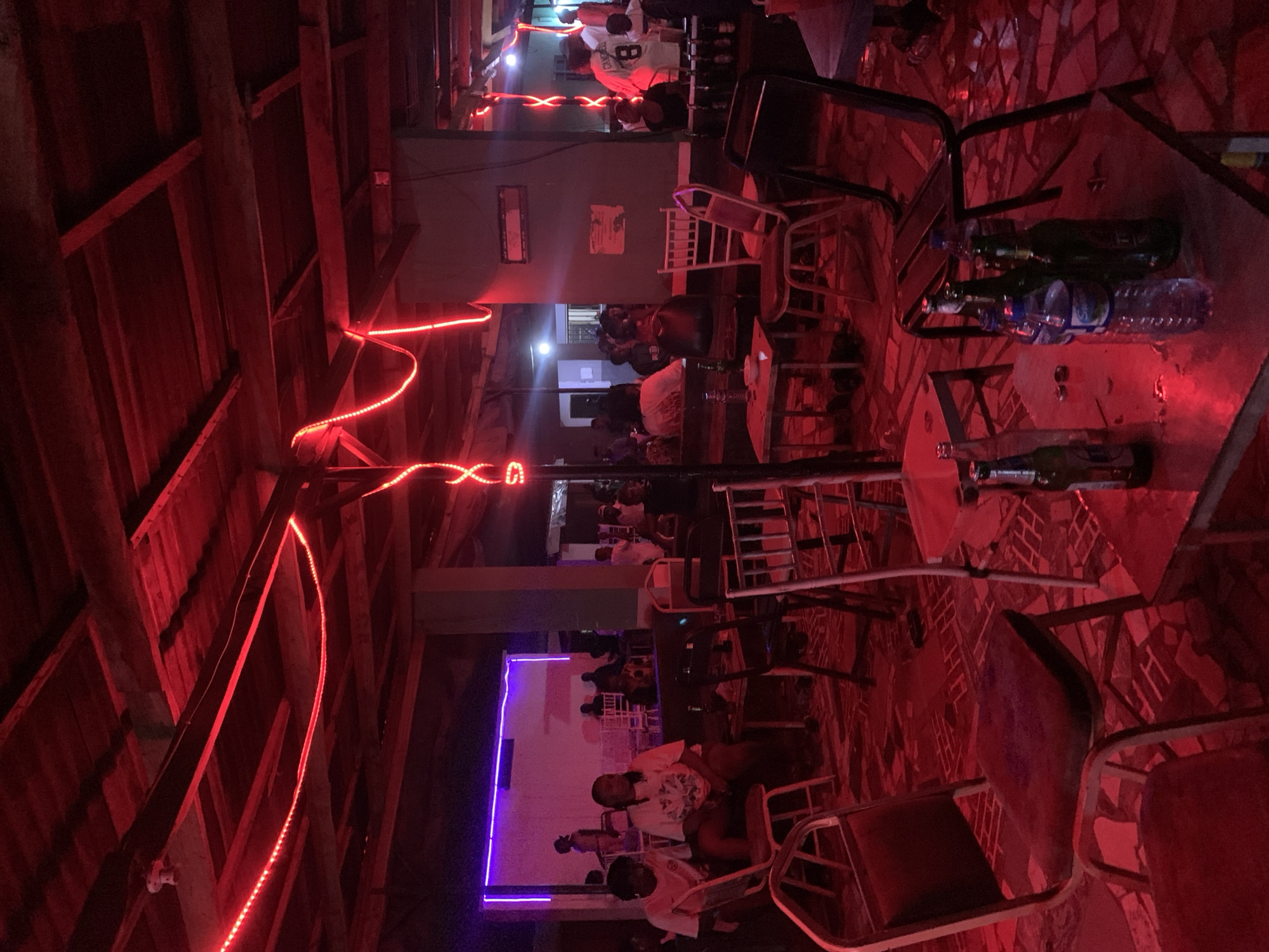 The image depicts the interior of a bar in Kabong, Nigeria. The space is illuminated by red neon lights strung along the wooden ceiling. Patrons are seated at tables, some with bottles of beer and water. People are casually dressed. The floor has a mosaic tile pattern. In the background, more patrons are visible, some in a room with purple lighting.