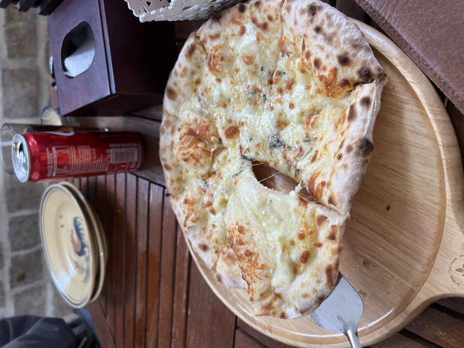 An overhead shot captures a freshly baked four-cheese pizza, likely Quattro Formaggi, served on a round wooden board placed on a slatted wooden table. The pizza is generously topped with various white cheeses, including visible pockets of blue cheese, melted and slightly browned in places, indicating a hot oven bake. Its crust is light and airy, with characteristic dark char spots. A metal pizza spatula is used to lift one slice, showcasing a substantial, gooey cheese pull.
To the left of the pizza, a red Coca-Cola can with an open tab rests on the table. Its label is partially visible, displaying the "Coca-Cola" logo and text like "ORIGINAL TASTE" and "TRADE MARK REGD." Below the can are two stacked, light-colored ceramic plates, the top one adorned with a stylized rooster design in blue and yellow. In the upper left background, a dark wooden tissue box with a carved pattern on top is visible. The slatted table and the paving visible beyond suggest an outdoor or semi-outdoor dining setting, consistent with a casual restaurant in Ninh Hòa, Vietnam, during the daytime. The overall scene depicts a casual meal being enjoyed. Faint, unreadable text or a logo is present on the lower right edge of the wooden pizza board.