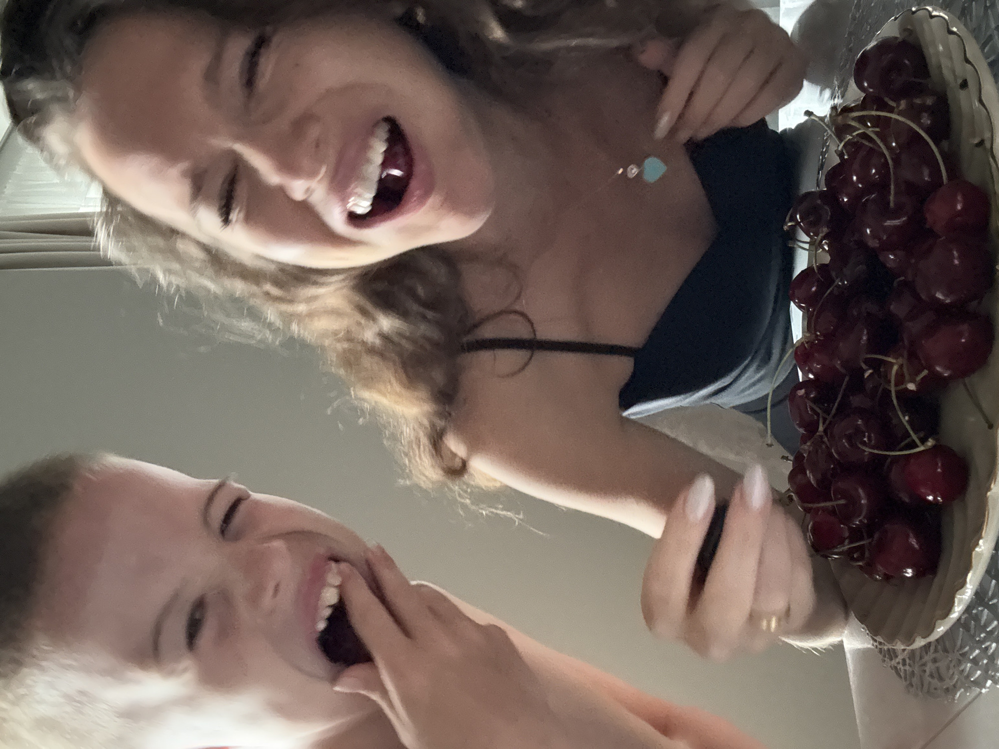 The image shows two young people, likely children or teenagers, close to the camera, with their faces filling the frame. The person in the foreground on the left has blonde hair and is laughing with their mouth wide open, their hand near their mouth. The person in the background on the right has brown curly hair and is also laughing exuberantly. They are wearing a dark top and a necklace with a light blue heart pendant. To the right of this person is a bowl filled with dark red cherries. The scene appears to be indoors, with a neutral-colored background that could be a wall or furniture. The lighting suggests it is daytime. Both individuals express joy and amusement. There is no visible text in the image. The location is Dolgoprudny, Russia.
