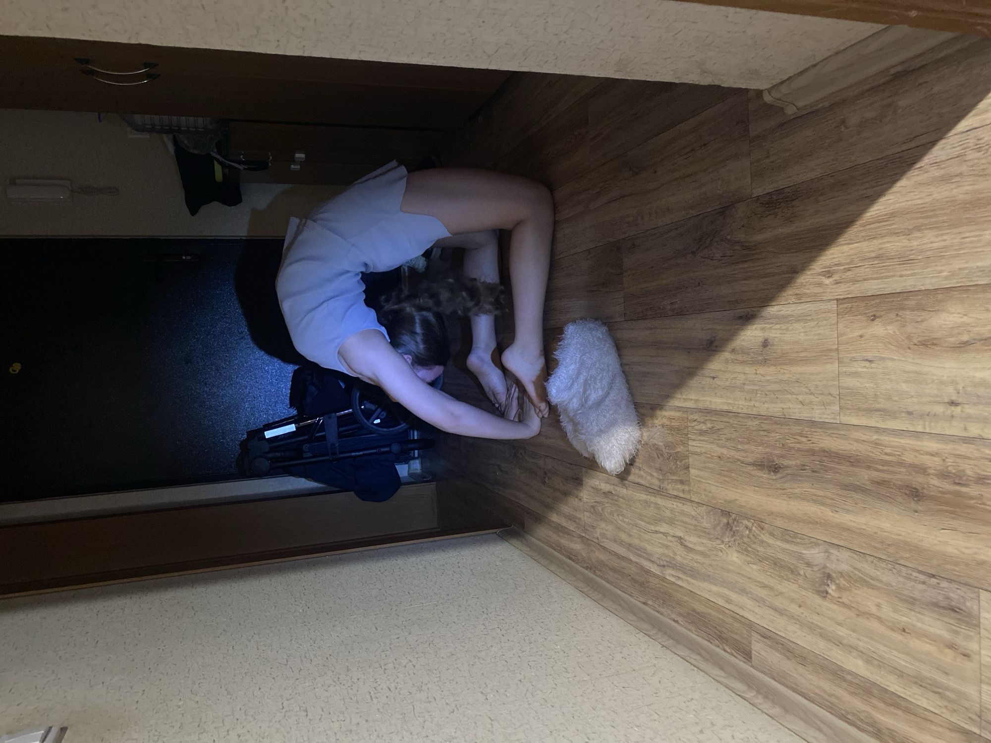 A high-angle indoor shot shows a young female in a contorted, arched-back yoga pose. She is wearing a light-colored shirt and shorts. Her hands appear to be touching her feet. Near her feet is a white, fluffy object, possibly a towel or rug. Next to her, to the left, is a folded stroller or baby carriage. The scene appears to be in a doorway or passageway between rooms. The floor is covered in light-colored wood-grain laminate. The lighting is subdued, with the light source above the subject. The setting location is the city of Unknown, Unknown.