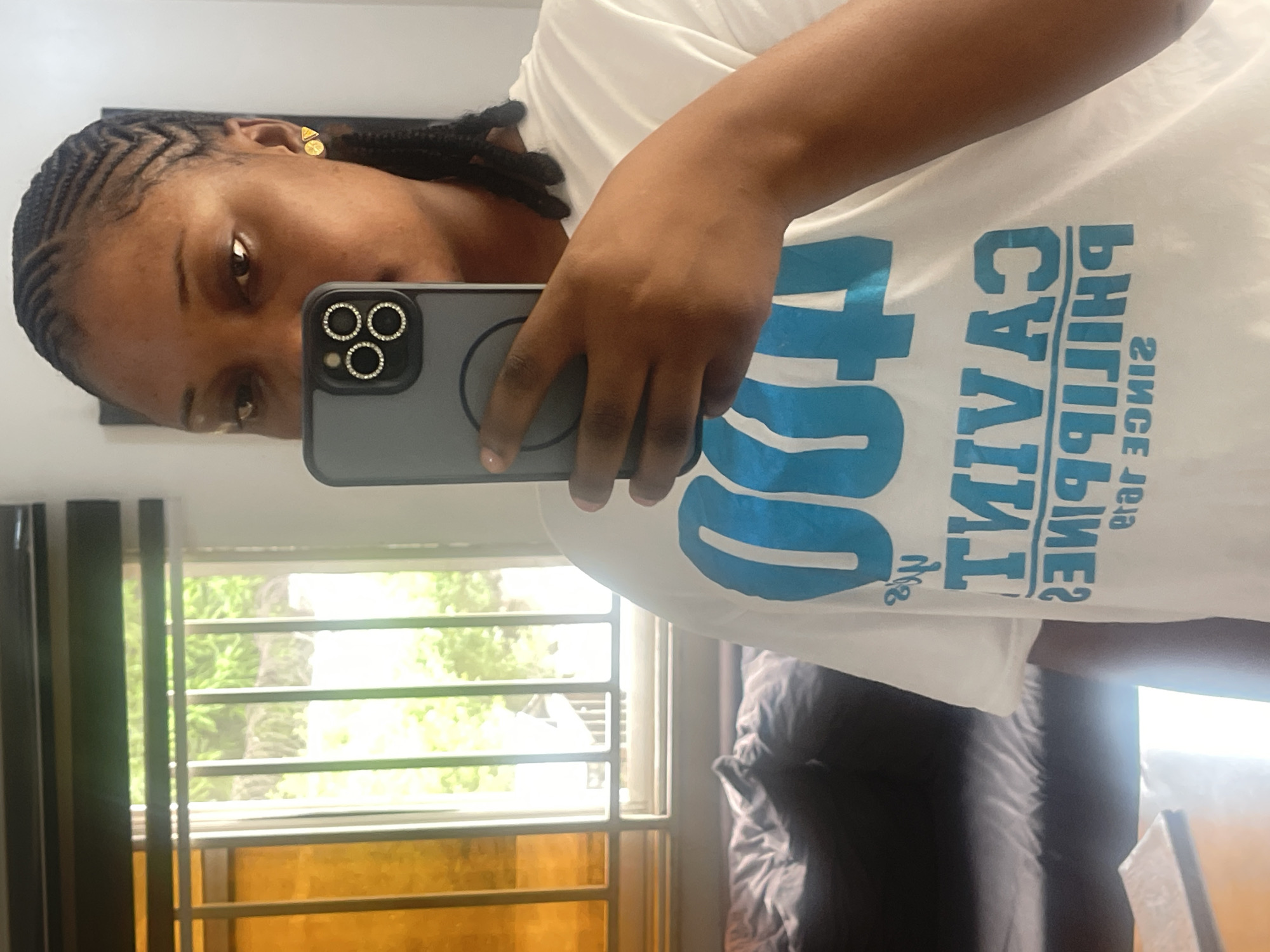 An individual is captured in a mirror selfie, holding a smartphone that partially obscures their face. The image is rotated ninety degrees to the left. The subject is a dark-skinned person, likely a woman, with dark hair styled in intricate cornrows along the hairline and braids further back. She wears small, gold-colored stud earrings. Her expression is neutral.
She is wearing a white t-shirt with prominent blue text printed on it. Due to the mirror reflection, the text appears reversed, reading "CAVITE PHILIPPINES SINCE 1898" (mirrored). Above "CAVITE", there's also mirrored text, possibly "PROVINCE OF".
The smartphone held by the subject has a dark-colored case embellished with clear rhinestones around the circular camera lenses. Her right hand, with light pink polished nails, holds the phone.
The setting appears to be indoors, with a window visible in the background, offering a glimpse of lush green foliage outside, suggesting a daytime scene in a residential or office environment. The window has white horizontal bars. To the left of the window, a dark vertical element, possibly a curtain or window frame, is present. A dark, possibly upholstered, object is also visible in the lower right background. This scene is located in Abuja, Nigeria.