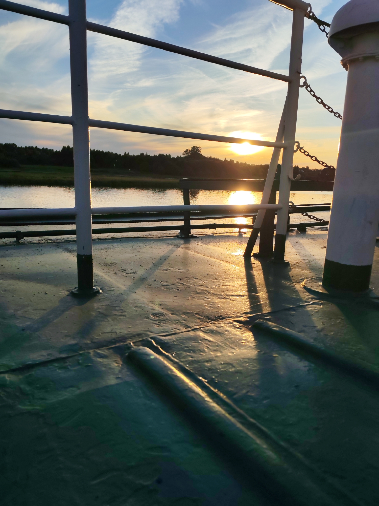 The image depicts a sunset scene viewed from the deck of a boat or ferry. The foreground shows the green painted deck with raised ribbed sections. The deck is wet, reflecting the warm, golden light of the setting sun. Shadows of metal railings and a mast-like structure are cast long across the wet surface, indicating the low angle of the sun.
In the mid-ground, a body of water, likely a river or lake, is visible. The water reflects the sky and the sun, creating a shimmering path of light. Beyond the water, a tree-lined shore is silhouetted against the bright sky.
The sky is a mix of blue and scattered clouds, with the sun positioned just above the horizon, casting its warm glow. The time of day is clearly sunset, characterized by the low, golden light and long shadows. The weather appears calm, with still water.
The visible objects include metal railings, chains, and a rounded structure resembling a mast or part of the boat's superstructure. The scene evokes a sense of tranquility and the beauty of a natural sunset.
The location context provided is Sheksna, Russia. There is no visible text in the image.