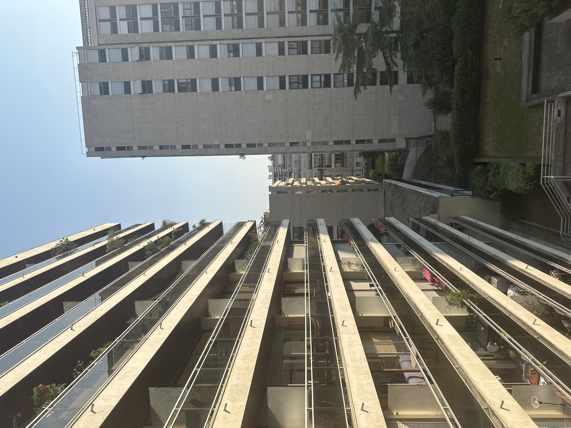 This is a vertical, eye-level, medium-wide shot looking up at two apartment buildings in Paris, France. The building on the left is a modern, multi-story building with numerous balconies, each featuring glass railings and beige concrete overhangs. Some balconies have plants and furniture visible. The building on the right is a taller, more austere concrete building with many uniformly spaced windows and white blinds. A section of this building on the far right is slightly further forward and shows more detailed window arrangements. In the foreground, to the right, is a patch of green lawn and trees, with what appears to be a paved courtyard area in the lower right corner. The sky is a clear, pale blue, indicating daytime. The overall scene depicts an urban residential environment.