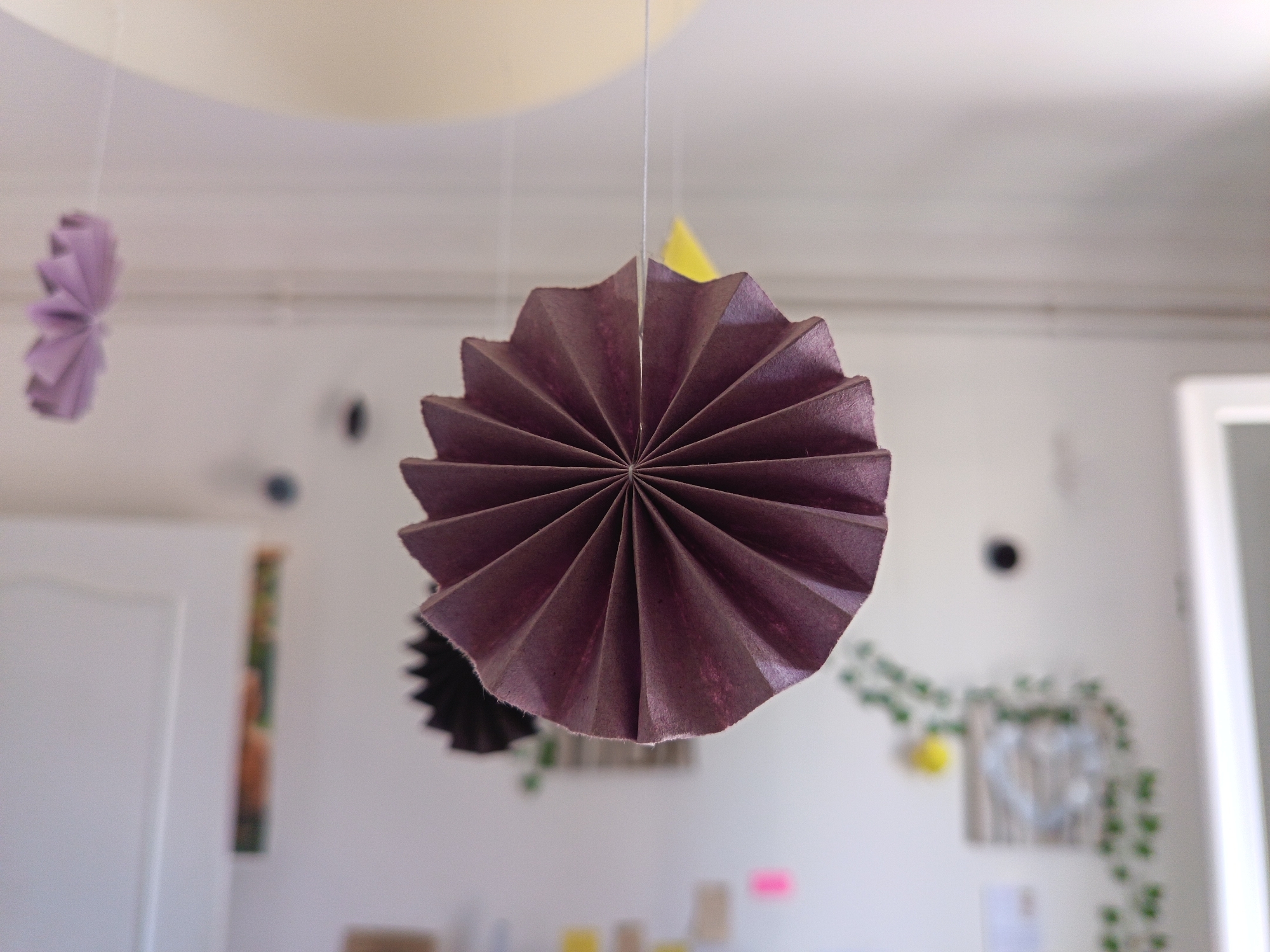 The image shows a close-up view of several folded paper decorations hanging from the ceiling in what appears to be a room. The primary focus is a large, dark purple fan-shaped decoration, centrally located and in sharp focus. To its left, a smaller, lighter purple fan-shaped decoration is visible, slightly out of focus. Behind the purple decorations, a yellow triangular decoration hangs. The background is a plain white wall with some unfocused elements, including a door on the left, some blurry green vine-like decorations with yellow circular objects, and some indistinct framed items. There are no people in the image. The overall impression is of an indoor decorative setting, possibly a child's room or a party space. The image does not contain any visual information that would allow for determining the location in the city of Périgueux, France.