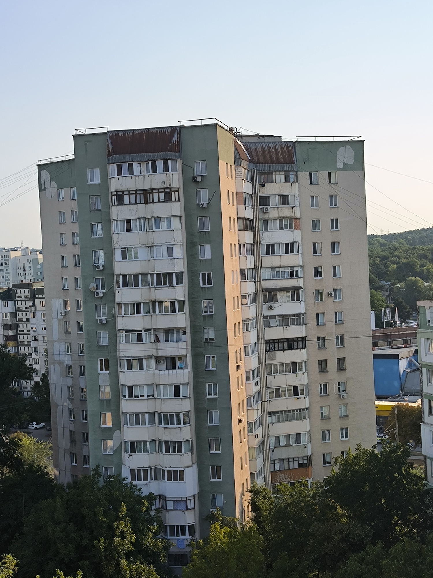 The image shows a high-rise apartment building in Chișinău, Moldova, with a clear sky and some surrounding greenery. The building is tall and appears to be constructed in sections with varying colors, primarily green and beige. It features numerous windows and balconies, some of which are enclosed. The top of the building has a slightly dilapidated, rusted metal roof structure. In the foreground, there are dense trees, suggesting the building is situated in a residential area with green spaces. The overall impression is of a typical urban residential complex in Eastern Europe.