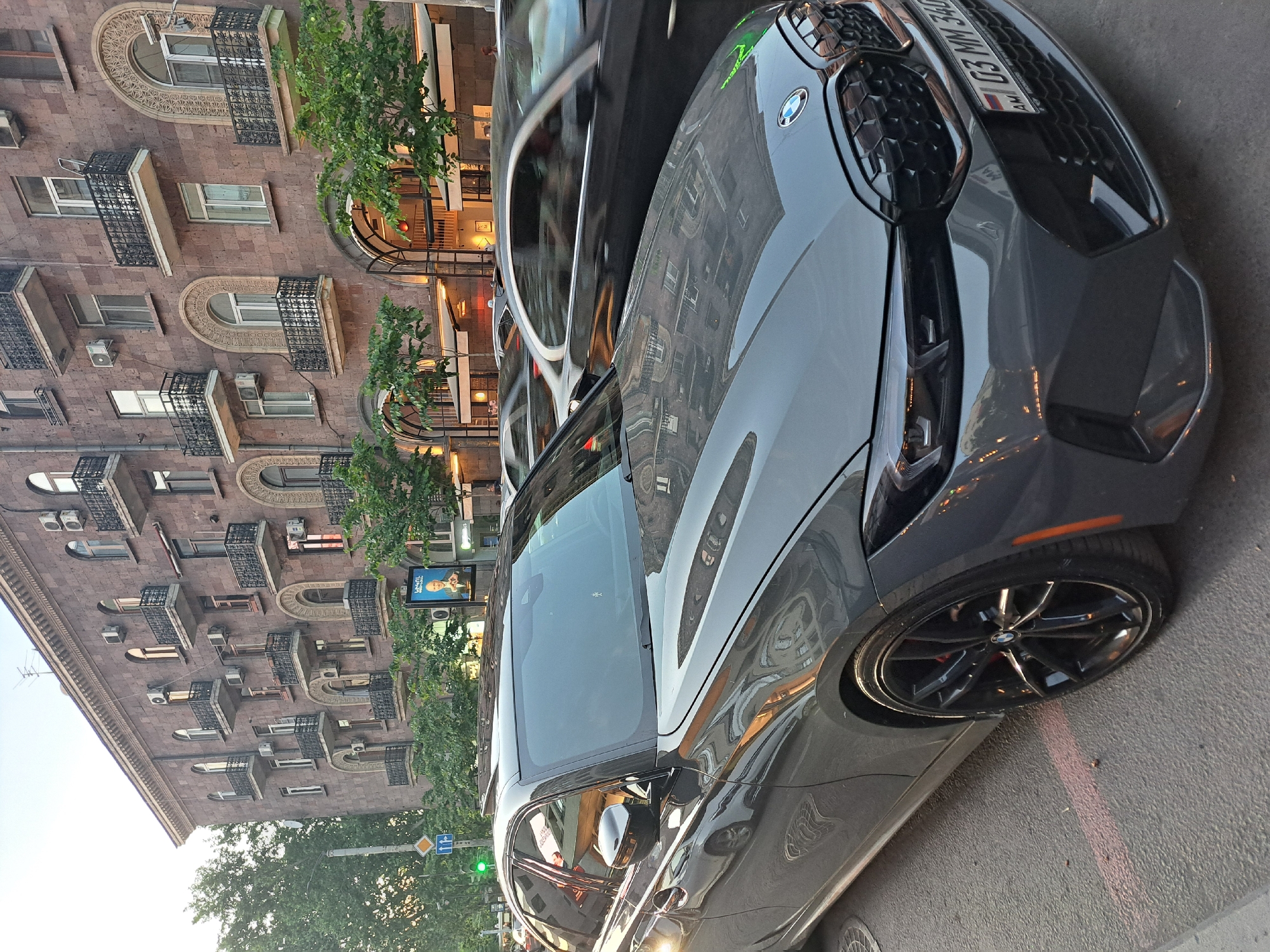 The image depicts a grey BMW parked on a street in Yerevan, Armenia. The car's front end is prominently displayed, revealing details like the headlight, the BMW emblem, and the Russian license plate 'G3 MM 340'. The reflection on the car's hood shows the surrounding architecture. Behind the car, a multi-story building with detailed arched windows and balconies is visible. Green trees line the street, and a street sign is present. The scene appears to be set during daylight, with the building illuminated by sunlight.